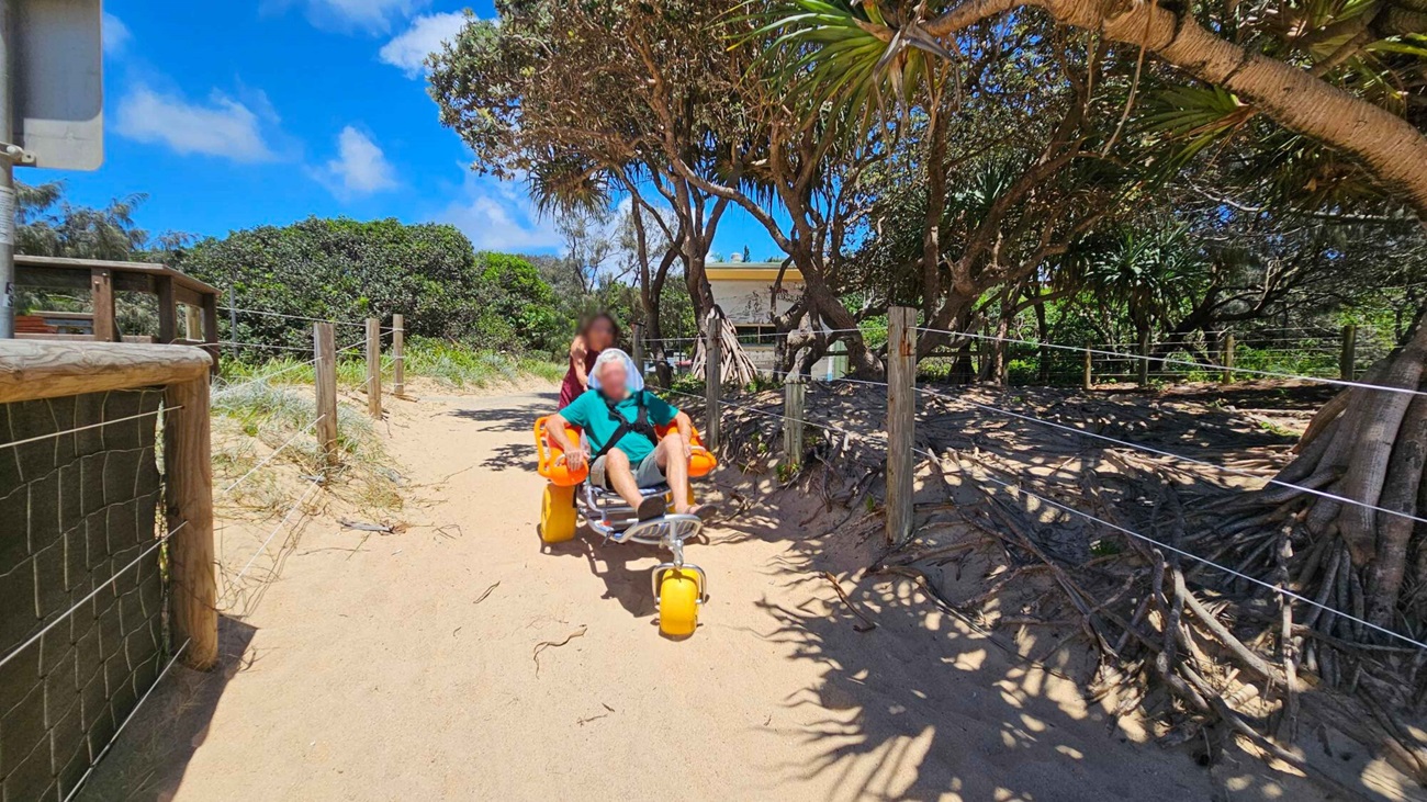 On the left, a wooden fence with wire mesh borders a sandy path lined with grass. On the right, trees with exposed roots and a wire fence run along the sand, casting dappled shadows. In the middle, a person sits in a yellow beach wheelchair with large inflatable wheels, pushed by another person. The foreground is sandy with uneven patches, and the background features dense green bushes and a building partially visible through the trees. On the left, a wooden fence with wire mesh borders a sandy path lined with grass. On the right, trees with exposed roots and a wire fence run along the sand, casting dappled shadows. In the middle, a person sits in a yellow beach wheelchair with large inflatable wheels, pushed by another person. The foreground is sandy with uneven patches, and the background features dense green bushes and a building partially visible through the trees.