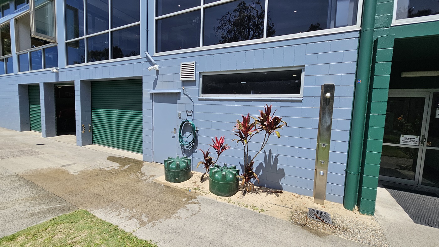 On the left, a green roller door with light blue walls and a small light blue door nearby. In the middle, a green hose coiled on the wall above two green cylindrical planters with red-leafed plants. On the right, a green-framed entrance leads into the building. The foreground has a damp concrete path bordered by grass, while the upper section features large windows reflecting trees. On the left, a green roller door with light blue walls and a small light blue door nearby. In the middle, a green hose coiled on the wall above two green cylindrical planters with red-leafed plants. On the right, a green-framed entrance leads into the building. The foreground has a damp concrete path bordered by grass, while the upper section features large windows reflecting trees.