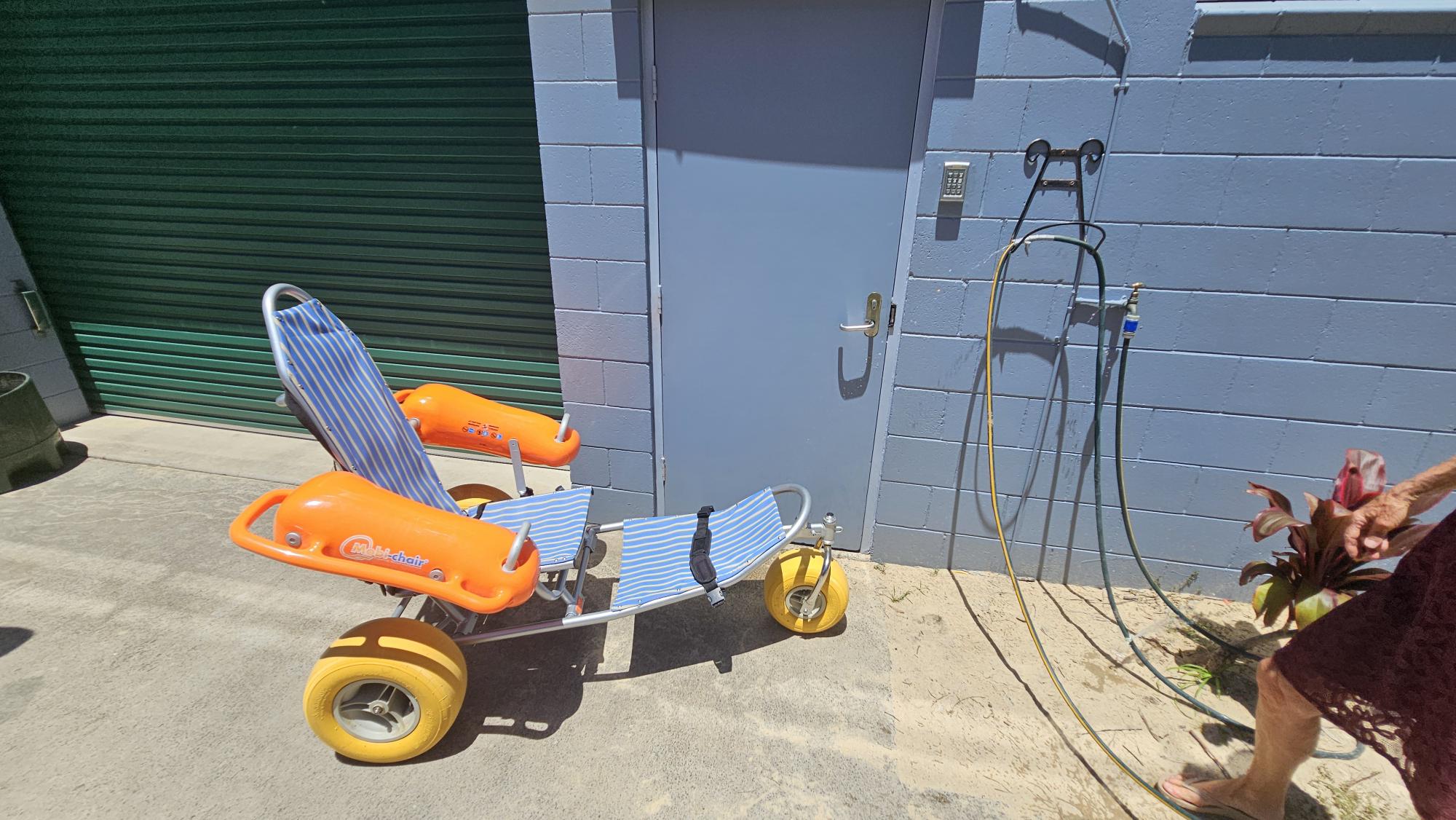 On the left, a green roller door set within a textured pale blue wall. On the right, a blue metal door with a hose attached to the wall beside it, featuring a showerhead and taps. In the foreground, a beach wheelchair with large yellow wheels, orange armrests, and a striped blue-and-white seat stands on a concrete floor transitioning into sand. Part of a person's arm and a plant with pinkish-red flowers are visible in the bottom right corner. There is no visible ceiling. On the left, a green roller door set within a textured pale blue wall. On the right, a blue metal door with a hose attached to the wall beside it, featuring a showerhead and taps. In the foreground, a beach wheelchair with large yellow wheels, orange armrests, and a striped blue-and-white seat stands on a concrete floor transitioning into sand. Part of a person's arm and a plant with pinkish-red flowers are visible in the bottom right corner. There is no visible ceiling.