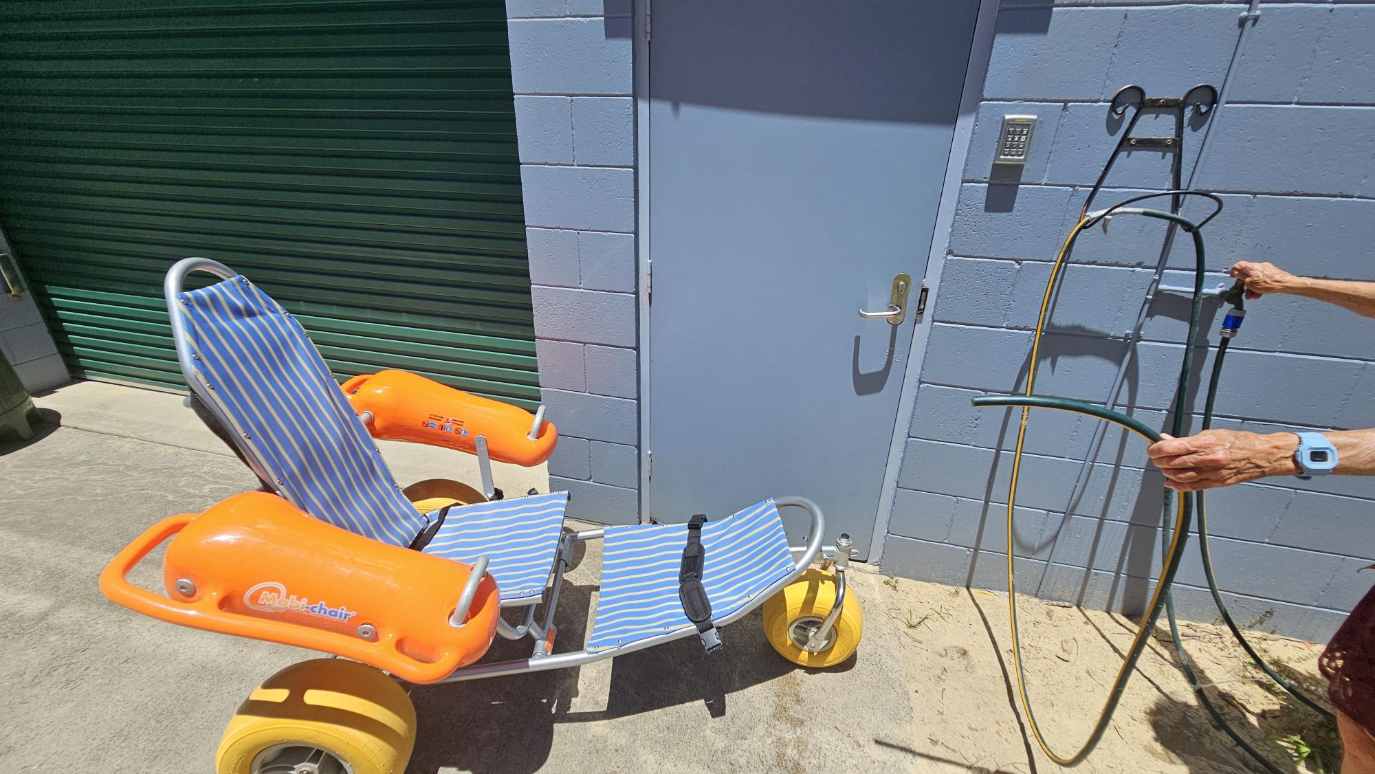 On the left, a green roller door set within a pale blue brick wall. On the right, a blue metal door with a silver handle is mounted on the same wall, next to a hose fixture with a showerhead attached. In the middle, a beach wheelchair with yellow wheels, orange armrests, and a blue-and-white striped seat is placed on a concrete surface. The foreground consists of concrete transitioning into sand, while a person's arm holding a green hose is visible on the right. No ceiling is present. On the left, a green roller door set within a pale blue brick wall. On the right, a blue metal door with a silver handle is mounted on the same wall, next to a hose fixture with a showerhead attached. In the middle, a beach wheelchair with yellow wheels, orange armrests, and a blue-and-white striped seat is placed on a concrete surface. The foreground consists of concrete transitioning into sand, while a person's arm holding a green hose is visible on the right. No ceiling is present.