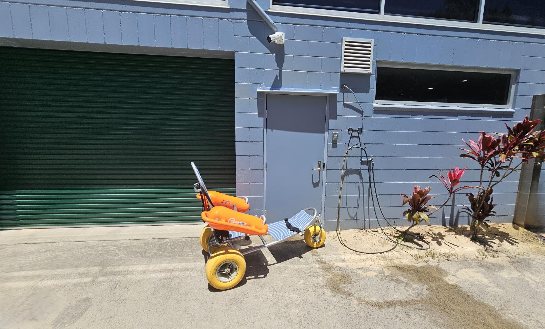 On the left, a closed green roller door set in a pale blue brick wall. On the right, a blue door with a silver handle, next to a wall-mounted black hose with a showerhead. In the middle, a yellow beach wheelchair with orange armrests and a blue-and-white striped seat rests on a concrete floor. The foreground consists of concrete with some sand closer to the right. In the background, red-leafed plants grow near the wall under a narrow horizontal window. Ceiling not visible. On the left, a closed green roller door set in a pale blue brick wall. On the right, a blue door with a silver handle, next to a wall-mounted black hose with a showerhead. In the middle, a yellow beach wheelchair with orange armrests and a blue-and-white striped seat rests on a concrete floor. The foreground consists of concrete with some sand closer to the right. In the background, red-leafed plants grow near the wall under a narrow horizontal window. Ceiling not visible.
