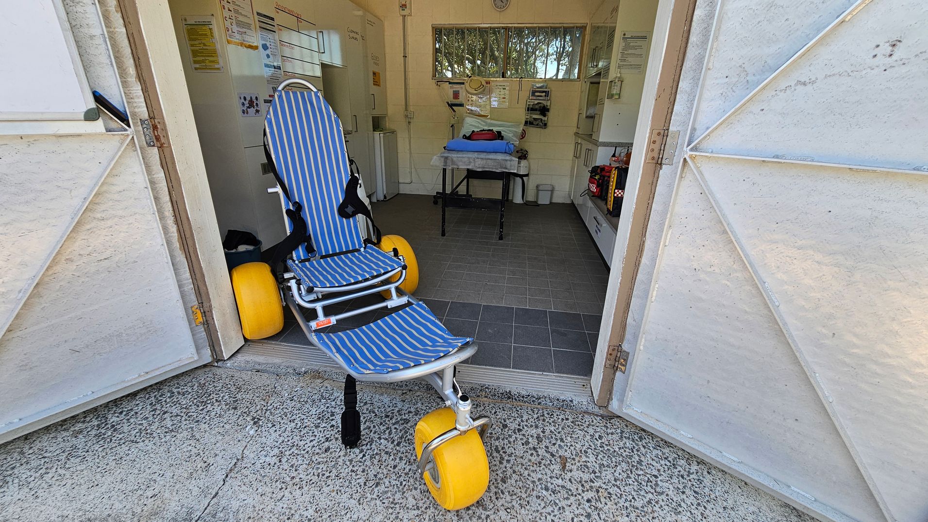 A room with open double doors leading to a tiled interior. In the foreground, a beach wheelchair with blue and white stripes and large yellow wheels is positioned at the entrance. Inside, a table with medical equipment is visible, along with various posters and notices on the walls. The floor is tiled, and there is a small window at the back with trees visible outside. The walls are light-coloured, and there are cabinets and shelves on the right. A room with open double doors leading to a tiled interior. In the foreground, a beach wheelchair with blue and white stripes and large yellow wheels is positioned at the entrance. Inside, a table with medical equipment is visible, along with various posters and notices on the walls. The floor is tiled, and there is a small window at the back with trees visible outside. The walls are light-coloured, and there are cabinets and shelves on the right.