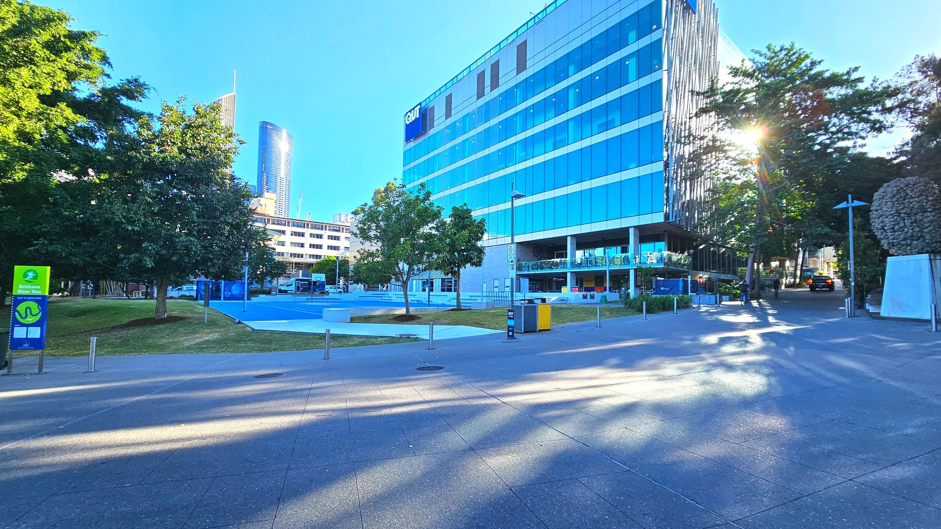 A modern urban scene featuring a large glass-fronted building with a sign reading 'QUT' on the upper floors. The building is surrounded by a paved area with scattered trees and a blue sports court. A sign for the Brisbane River Run is visible on the left. The area is open with bollards lining the pathways. A sculpture resembling a cluster of spheres is on the right, and a few people are walking nearby.