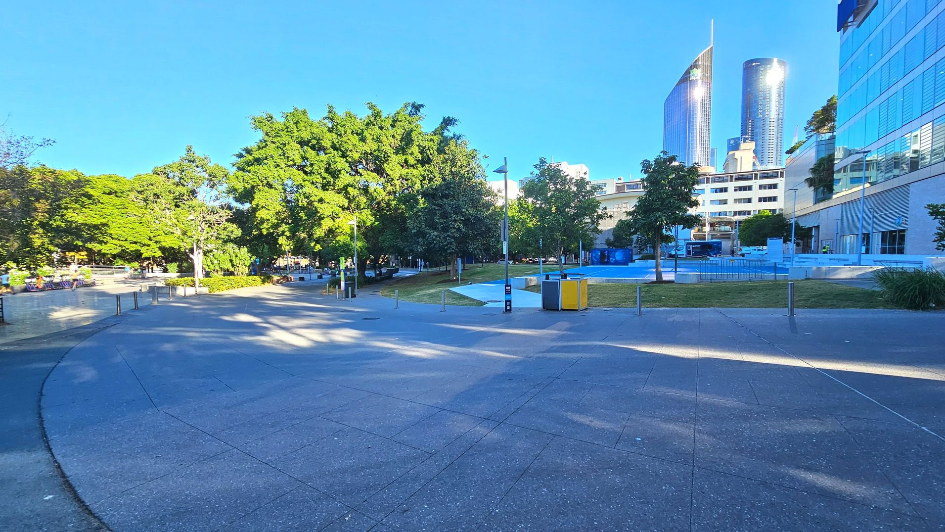 A spacious urban area with a wide, curved concrete path leading through a park-like setting. On the left, lush green trees provide shade over benches and a low wall. To the right, modern glass buildings rise, reflecting light. A small grassy area with scattered trees and a few bollards separates the path from the buildings. A lamppost and a pair of yellow and grey bins are visible near the centre.