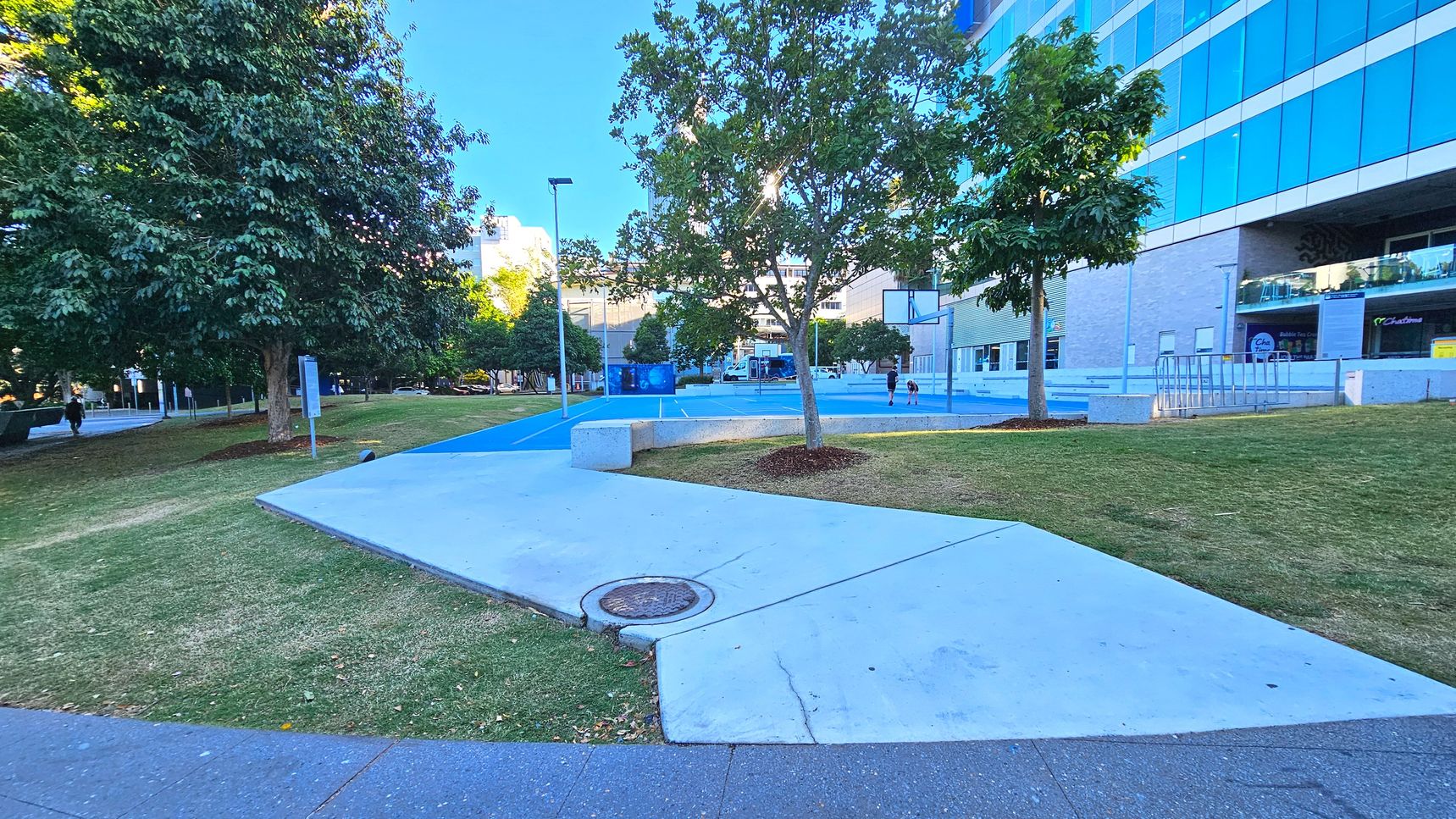 A modern outdoor area featuring a blue basketball court with a hoop, surrounded by grass and concrete paths. A large building with reflective glass windows is on the right, and several trees provide shade. A concrete bench is near the court, and a bike rack is visible against the building. The area is open and spacious, with a mix of natural and urban elements.
