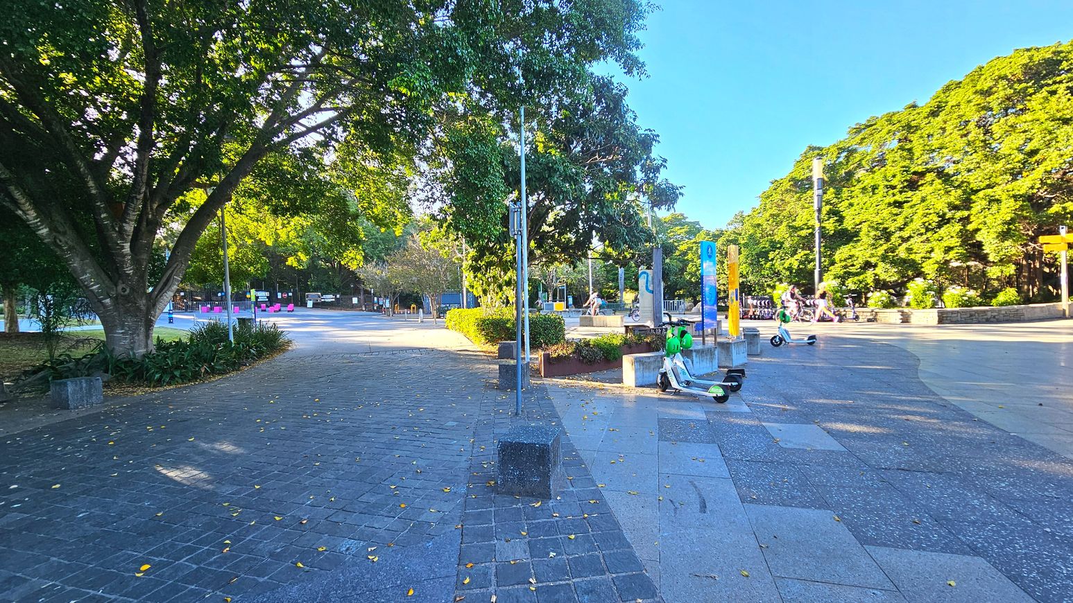 A spacious outdoor area with a mix of paved surfaces, including dark grey cobblestones and lighter concrete tiles. Large trees with dense foliage provide shade on the left, while the right side is more open. Several electric scooters are parked near a concrete block. In the background, pink chairs and signage are visible, suggesting a recreational space. The area feels open with clear pathways for walking.