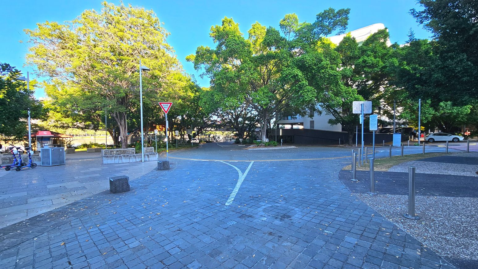 A paved area with a 'Give Way' sign on a pole in the centre, surrounded by large leafy trees. The ground is covered with grey and blue square tiles, transitioning to a pebbled surface on the right. Metal bollards line the edge of the path. A small kiosk with a red roof is visible to the left, and a white car is parked on the right. The space feels open with trees providing a natural boundary.