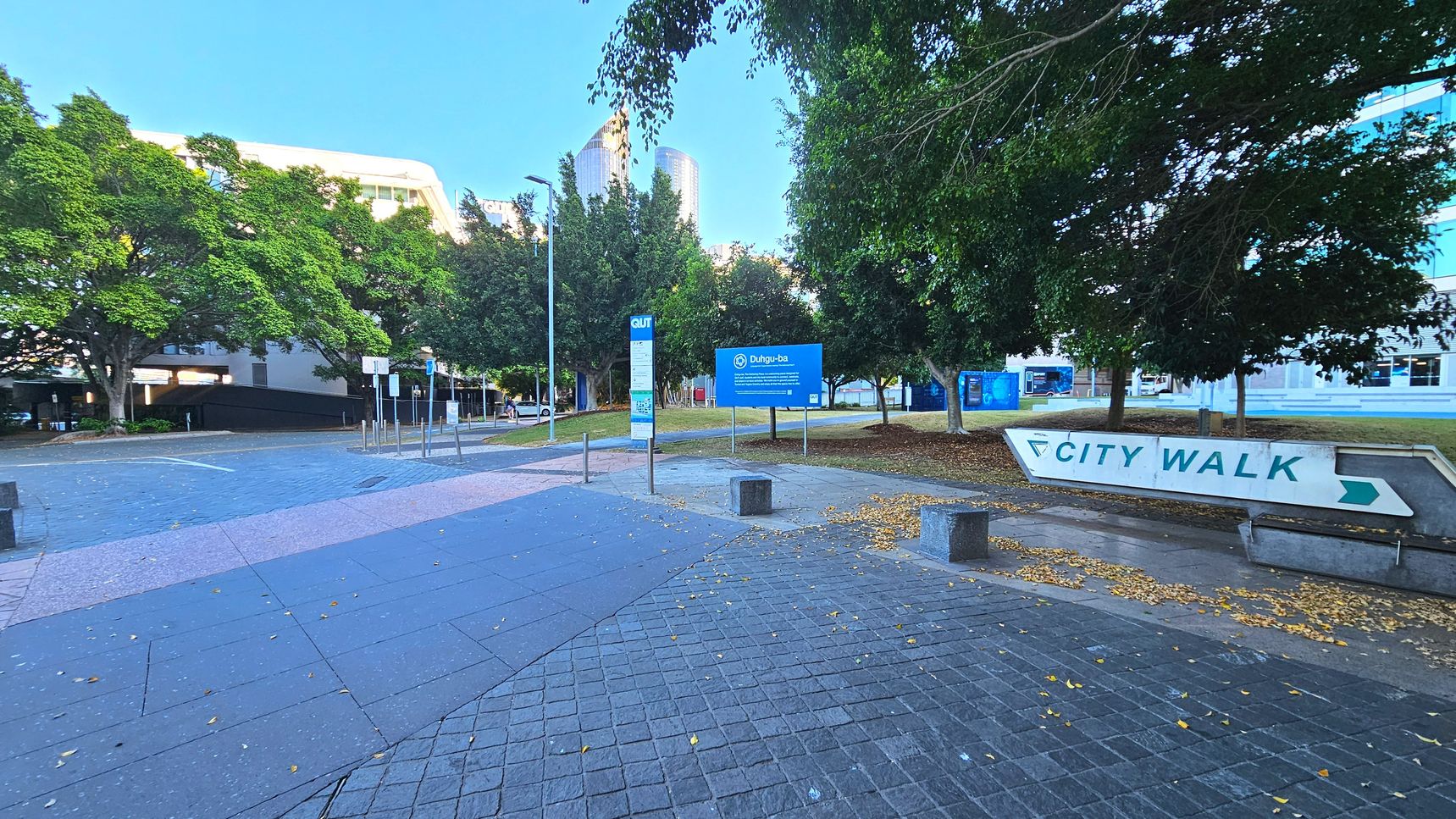 A paved area with a large 'City Walk' sign pointing right is visible. The ground is covered with grey and pink tiles, transitioning to a cobblestone pattern. Several trees with dense foliage line the area, providing shade. A blue sign with white text stands near the centre, and a tall building is partially visible in the background. Metal bollards and a lamppost are also present, indicating a pedestrian-friendly zone.
