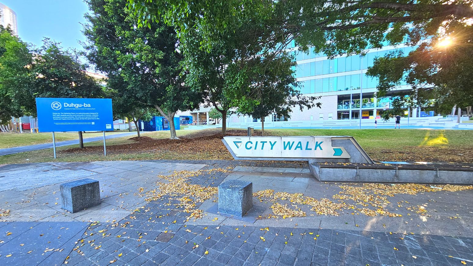 A paved area with a large white sign reading 'CITY WALK' in blue letters and an arrow pointing right. The sign is set on a low concrete structure. Nearby, a blue sign with 'Duhgu-ba' is visible, mounted on metal posts. The area is bordered by trees with scattered fallen leaves on the ground. In the background, a modern glass-fronted building is visible, with steps leading up to it.