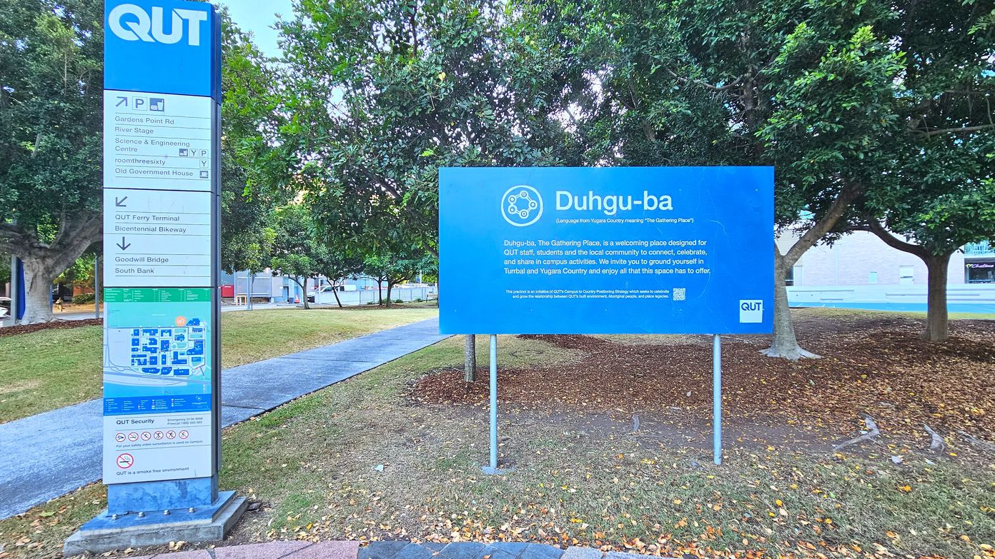 A large blue sign with white text stands on a grassy area, reading 'Duhgu-ba' and describing it as a gathering place. To the left, a tall directional sign with the QUT logo lists nearby locations and includes a campus map. The area is bordered by trees, with a paved path running through the grass. The ground is covered with fallen leaves, and a building is partially visible in the background.