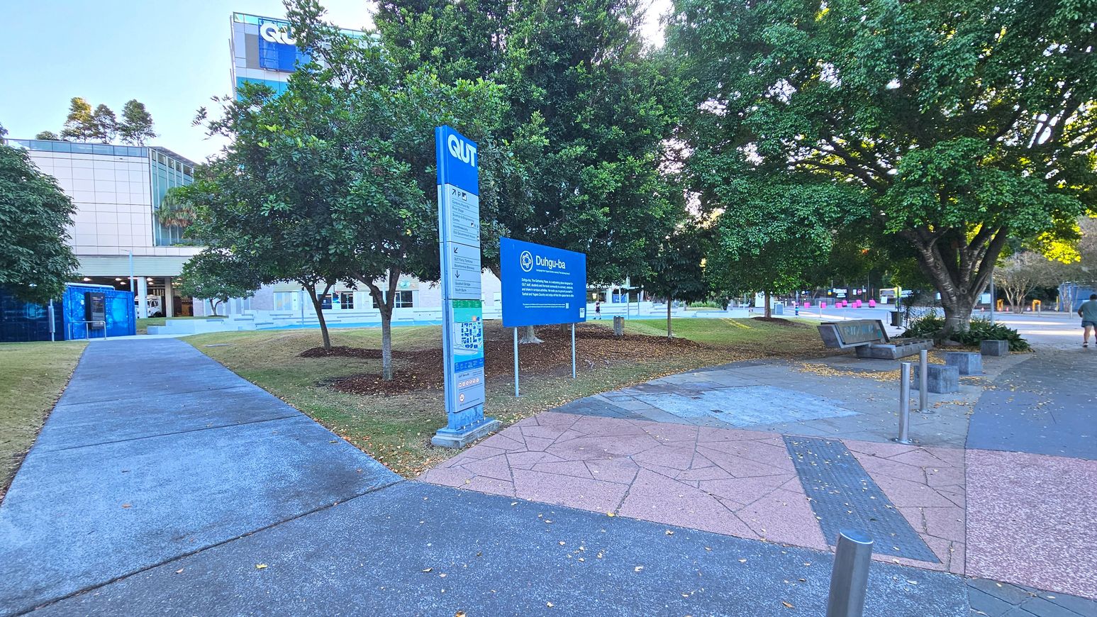 A campus scene with a prominent blue sign reading 'Duhig-bah' near a tall directory sign marked 'QUT'. The area features a mix of concrete and pinkish stone pathways, bordered by grass and several large trees. A modern building with glass and white panels is visible in the background. There are benches and bollards along the path, and a person walking in the distance.