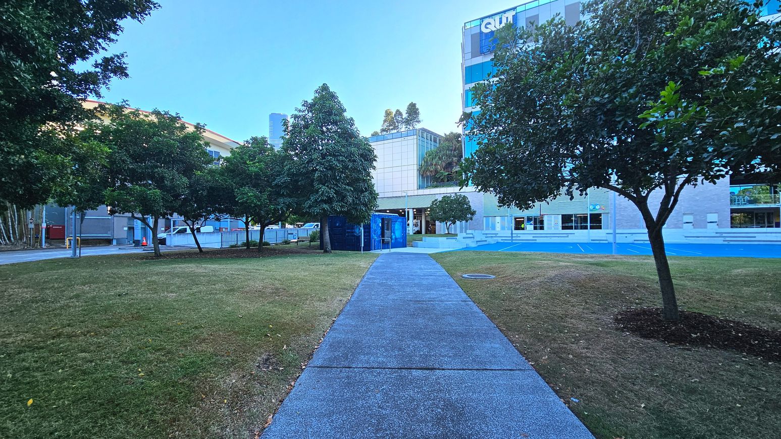 A paved path leads through a grassy area with trees on either side, towards a modern building with large glass windows. The building has a sign with the letters 'QUT' visible. To the left, a blue structure is partially obscured by trees. The area is open with no visible barriers, and the path is bordered by grass transitioning to a concrete surface near the building.