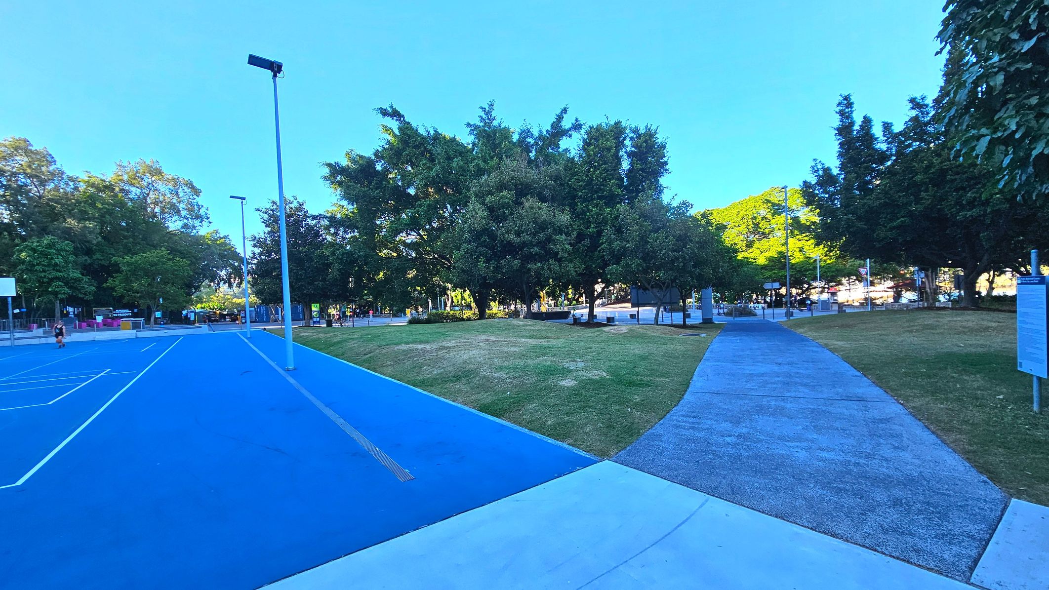 A vibrant outdoor scene featuring a blue sports court on the left with white lines and a basketball hoop. Adjacent to the court is a grey concrete path curving through a grassy area with scattered trees. Tall streetlights line the court and path. In the background, there are more trees and some park signage. The area appears open and spacious, with a mix of natural and man-made elements.