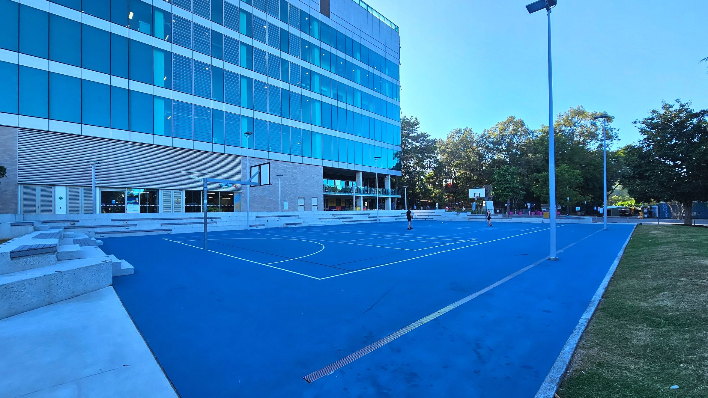 A modern outdoor basketball court with a bright blue surface is adjacent to a multi-storey building with large glass windows and horizontal louvres. The court features two basketball hoops and is surrounded by tall street lamps. Concrete seating lines the edge of the court, and a grassy area borders one side. Trees are visible in the background, providing a natural boundary.