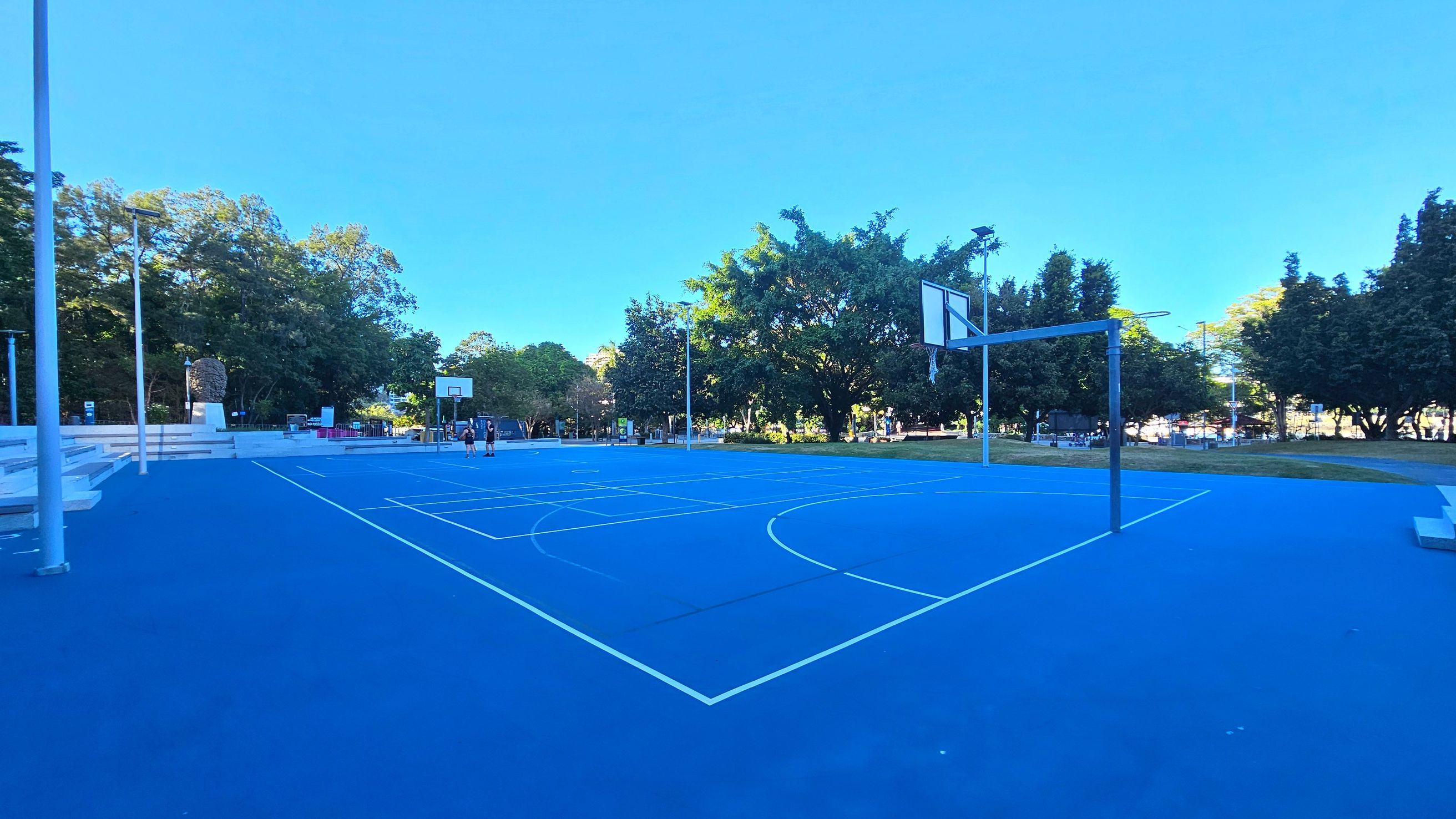 A vibrant blue outdoor basketball court with white lines is surrounded by lush green trees. Two basketball hoops are visible, one in the foreground and another in the distance. To the left, there are tiered concrete seating steps. Several tall light poles are positioned around the court. The area is open and bordered by grass and trees, providing a natural enclosure.