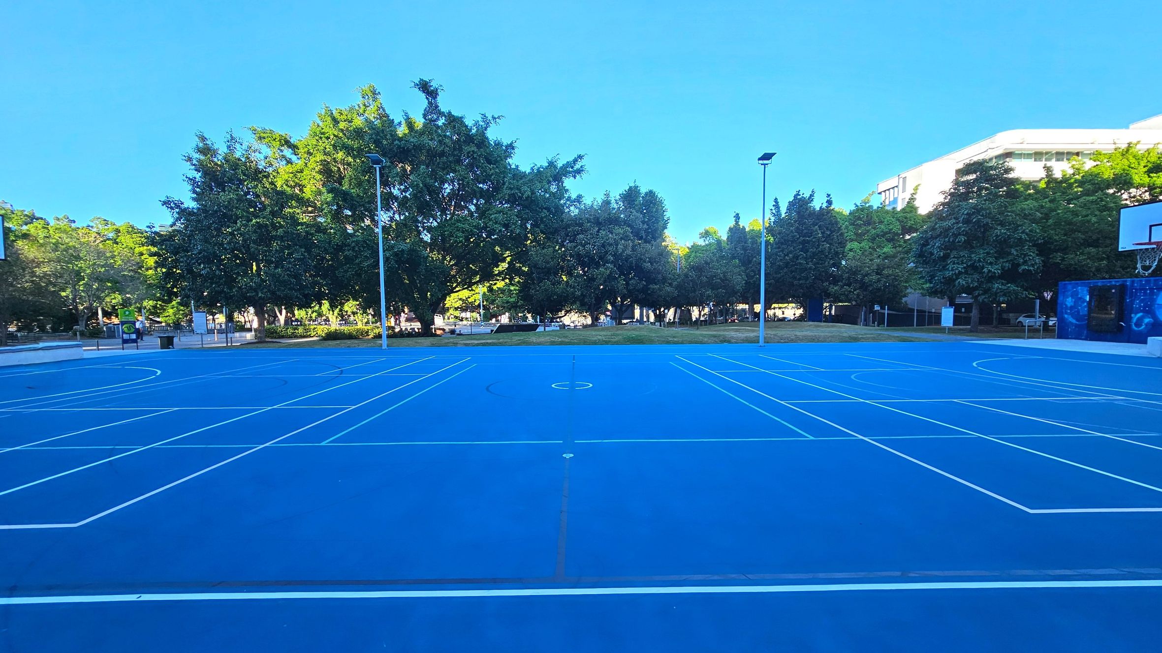 A vibrant blue outdoor basketball court with white lines is centred in the image. Two tall streetlights are positioned on either side of the court. Surrounding the court are lush green trees and a grassy area. To the right, a basketball hoop is visible, and a building peeks through the trees. The court is open and expansive, with no visible barriers, providing a clear view of the surrounding greenery.