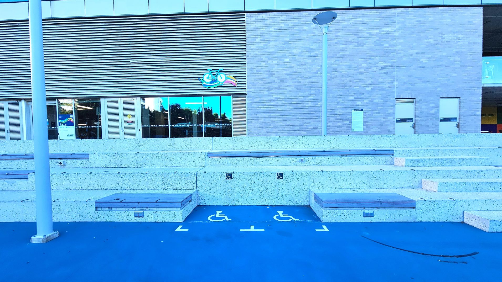 The image shows a modern building facade with a combination of brick and metal louvre panels. In front, there are tiered concrete seating steps with wooden tops, flanked by two designated wheelchair spaces marked on a blue surface. A tall lamp post stands to the left. The building features large glass doors and windows, with a colourful logo above. The area feels open with no visible barriers.