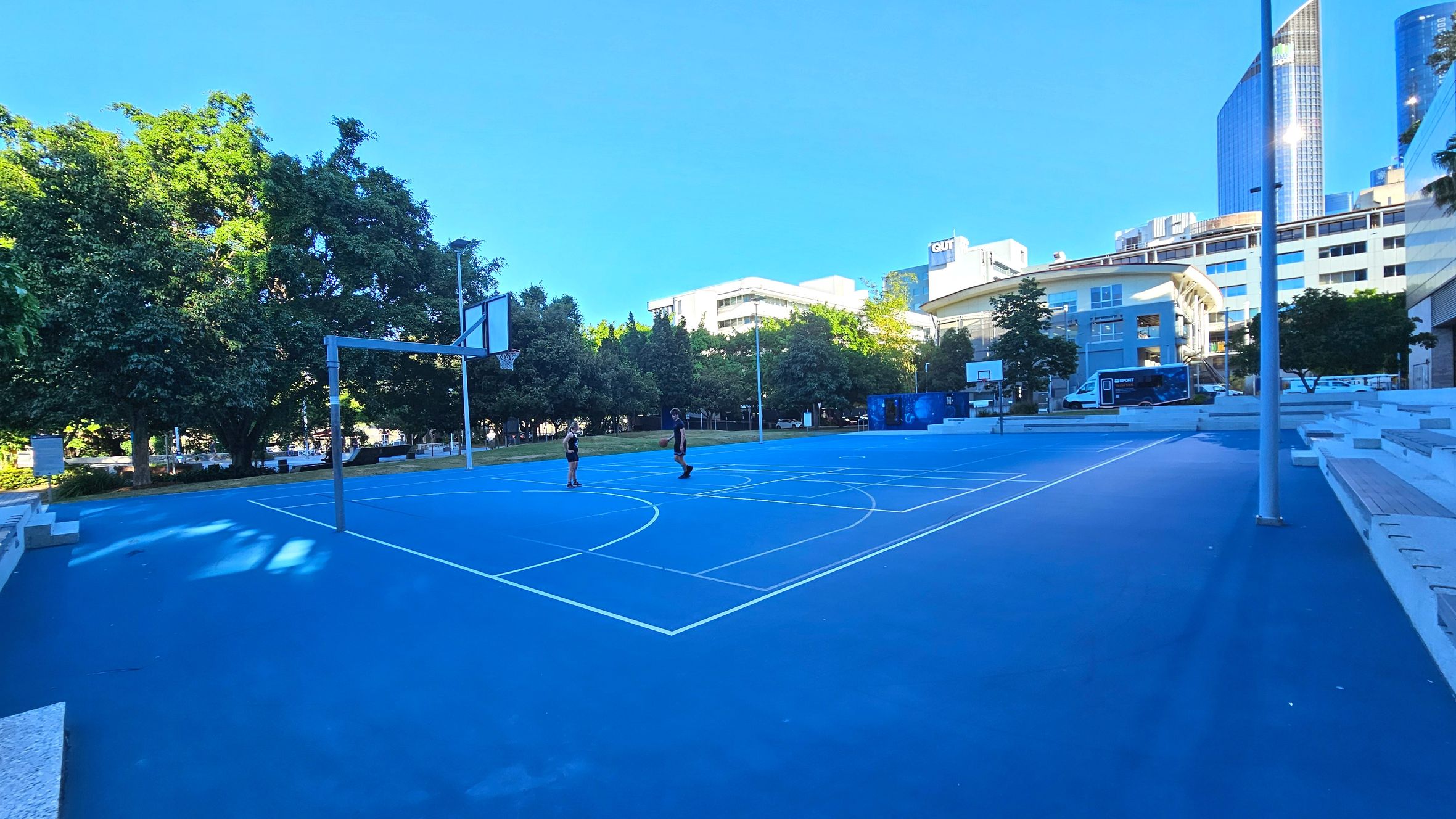 A vibrant blue outdoor basketball court with white lines is surrounded by trees and modern buildings. Two people are playing on the court. The court is bordered by tiered concrete seating on one side. A tall building with reflective glass and a sign reading 'QUT' is visible in the background. The area is open with trees providing some shade, and a van is parked nearby.