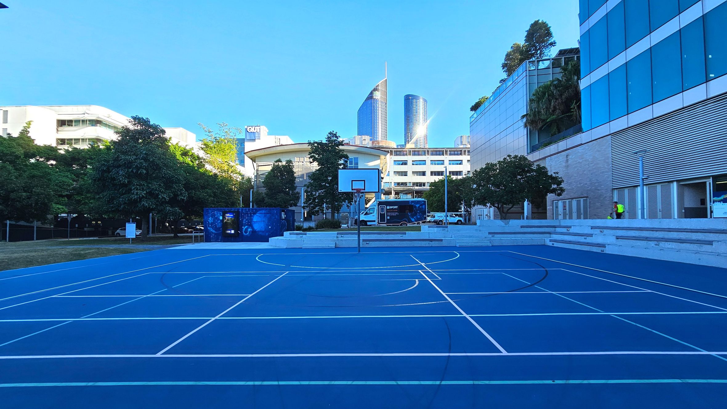 A vibrant blue basketball court with white lines is centrally located, surrounded by modern buildings. A basketball hoop stands prominently in the middle. To the right, a glass-fronted building with greenery on the roof is visible. In the background, tall skyscrapers rise, reflecting light. Trees and a small grassy area are on the left, while tiered concrete seating lines the right side.