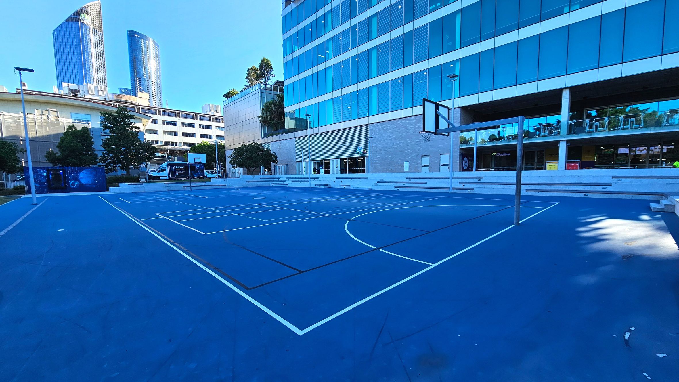 A blue outdoor basketball court with white lines is situated in front of a modern glass building. The building has multiple floors with large windows and a balcony with tables and chairs. Two basketball hoops are visible, one at each end of the court. To the left, there are trees and a smaller building. The area is open with no visible barriers, and the court surface is smooth and even.