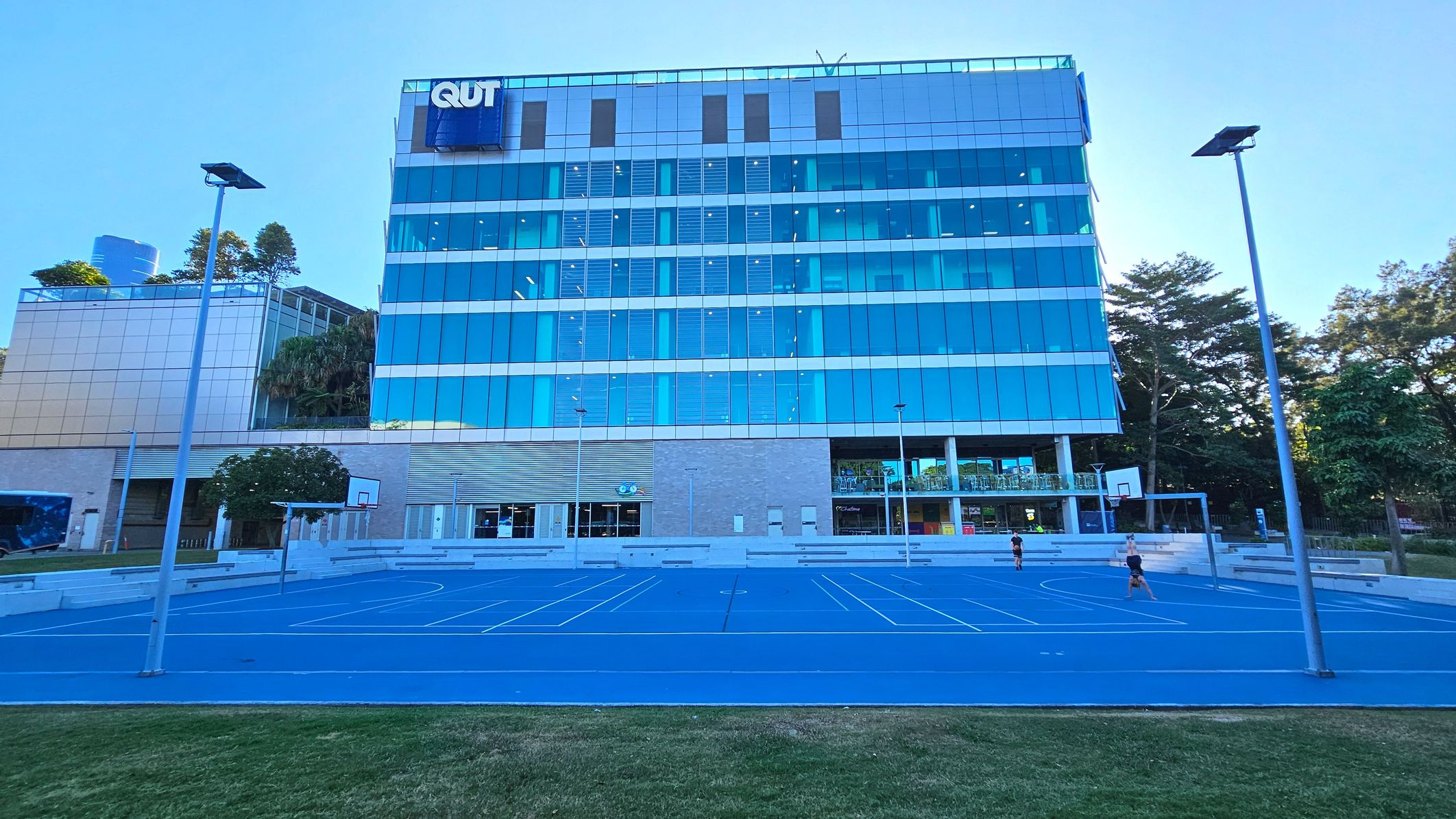A modern multi-storey building with a glass facade, featuring the letters 'QUT' at the top. In front of the building is a blue sports court with basketball hoops and white line markings. The court is surrounded by low concrete seating and bordered by grass. Tall street lamps line the court, and trees are visible to the right. The building has a mix of glass and brick surfaces, with a covered entrance area.