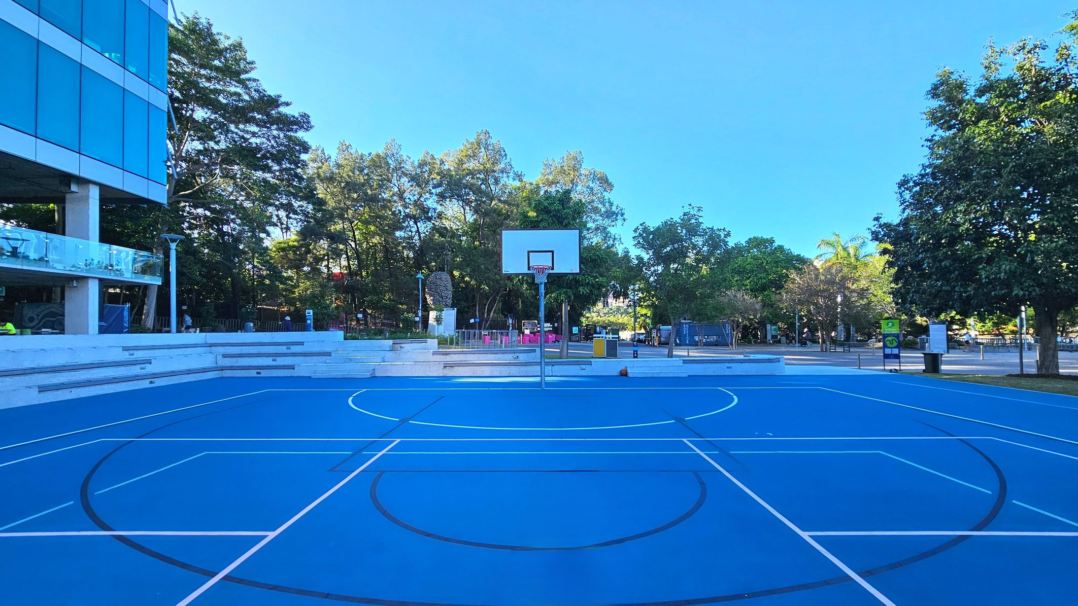 A vibrant blue basketball court with white and black lines is centrally positioned. A basketball hoop stands at the far end. To the left, a modern building with large glass windows and a balcony overlooks the court. Surrounding the court are tiered concrete seating steps. Trees and greenery form a natural boundary in the background, with various signs and a bin visible near the seating area.