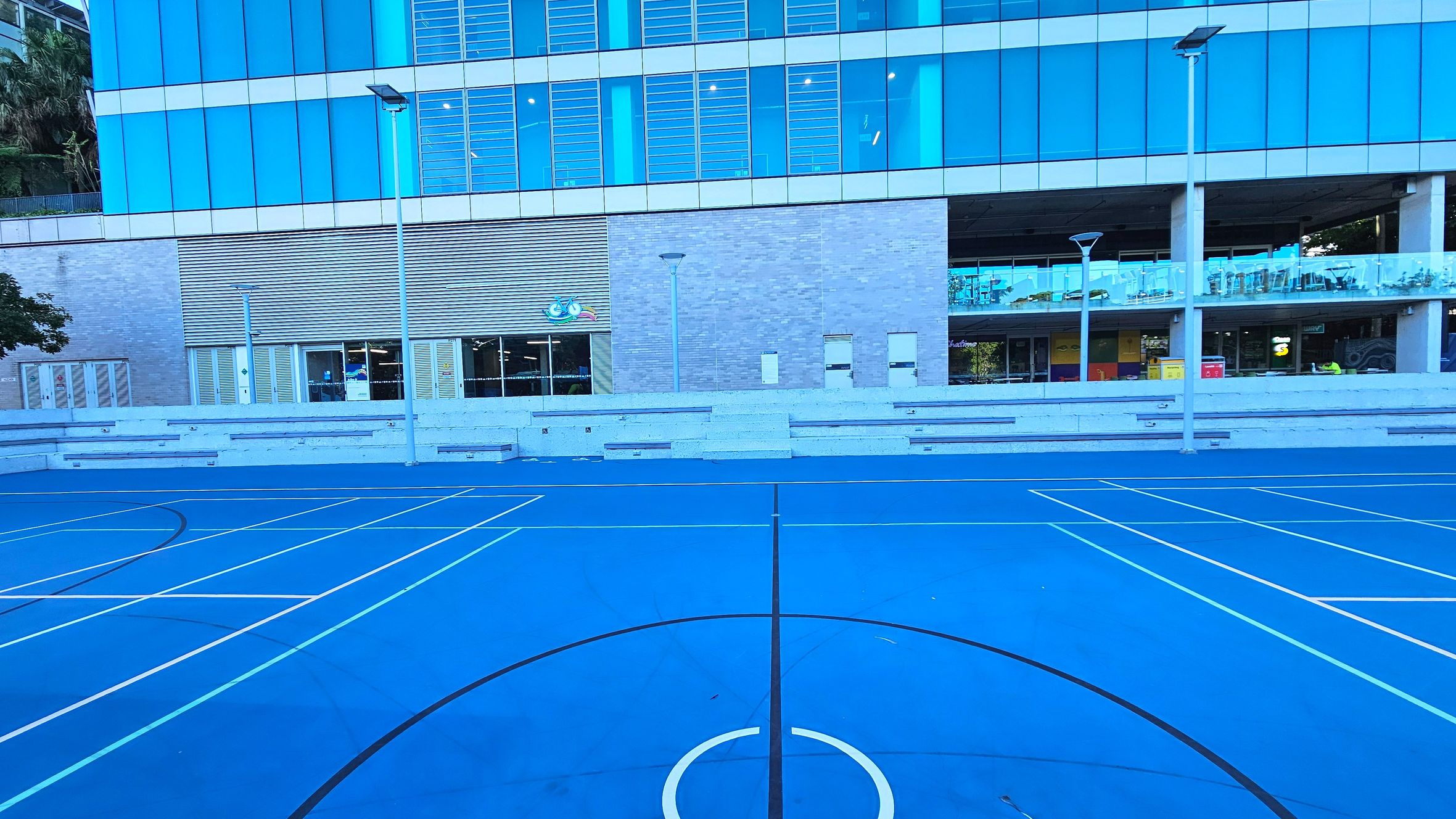 A vibrant blue sports court with white lines for various games is in the foreground. Behind it, a modern building with large blue-tinted glass windows and a grey brick facade stands. The building features a covered walkway with glass railings and several entrances. Tall street lamps line the court, and tiered concrete seating is visible along the building's base.