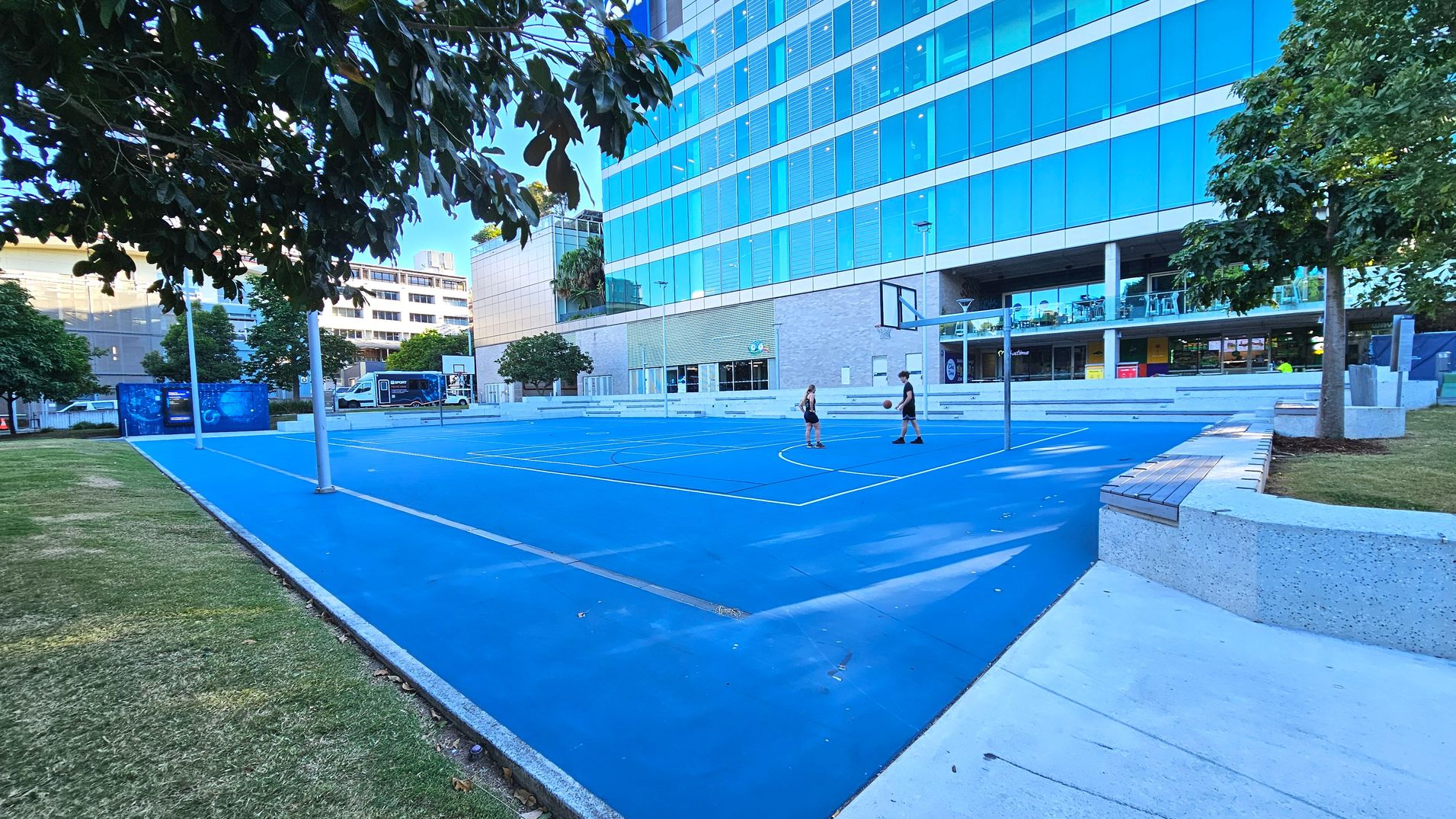 A vibrant blue basketball court is situated next to a modern glass building with reflective windows. Two people are playing basketball on the court, which is surrounded by a low concrete wall with wooden seating. Trees with lush green foliage border the court, and a grassy area is visible to the left. The building features a ground-level entrance and a balcony with seating, suggesting a recreational or commercial space.