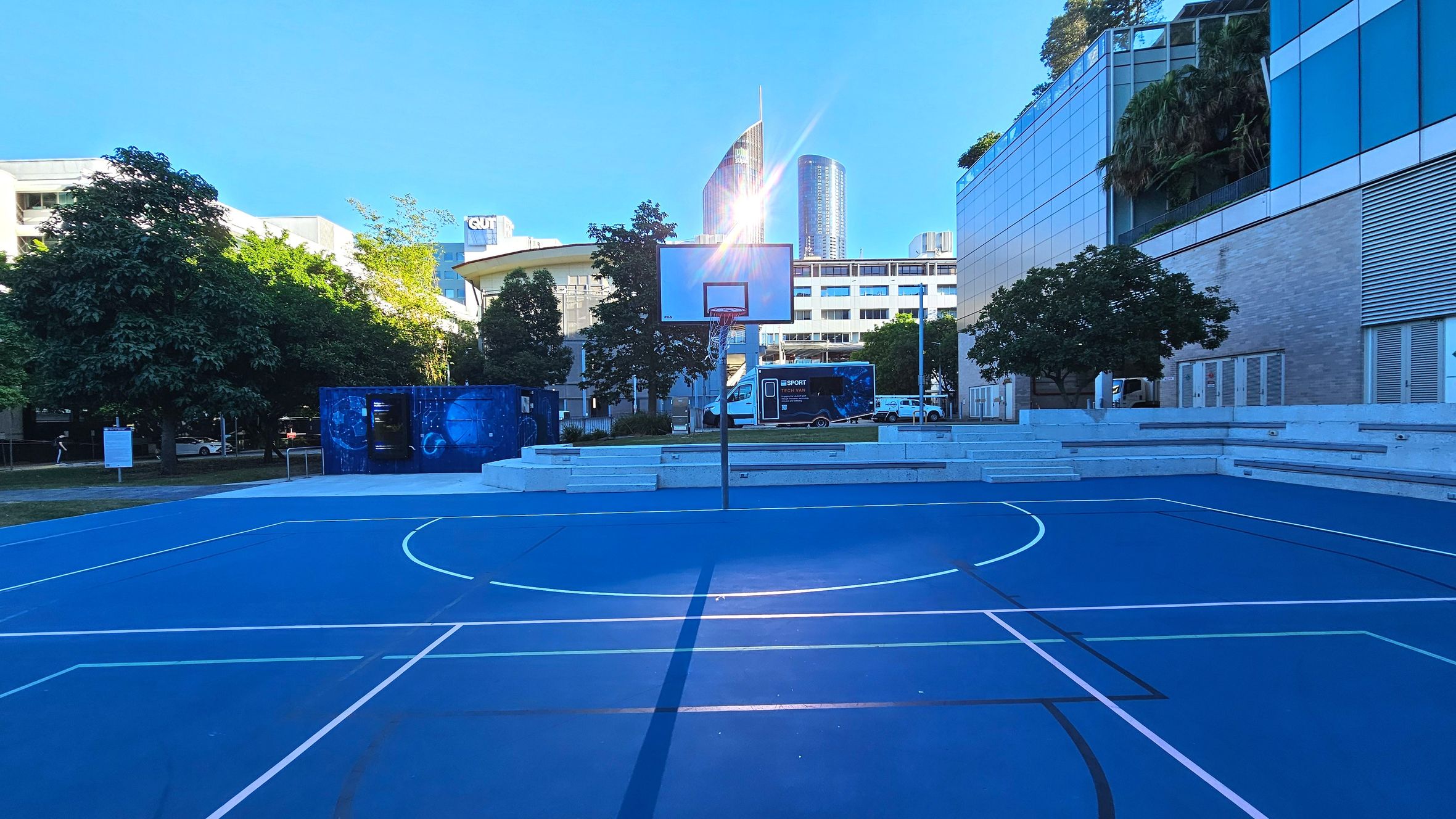 A blue outdoor basketball court with white lines is surrounded by tiered concrete seating on the right. A basketball hoop stands centrally on the court. To the left, trees and a blue structure with circular designs are visible. In the background, modern buildings with glass facades rise, including a curved tower. The area feels open with a mix of natural and urban elements.