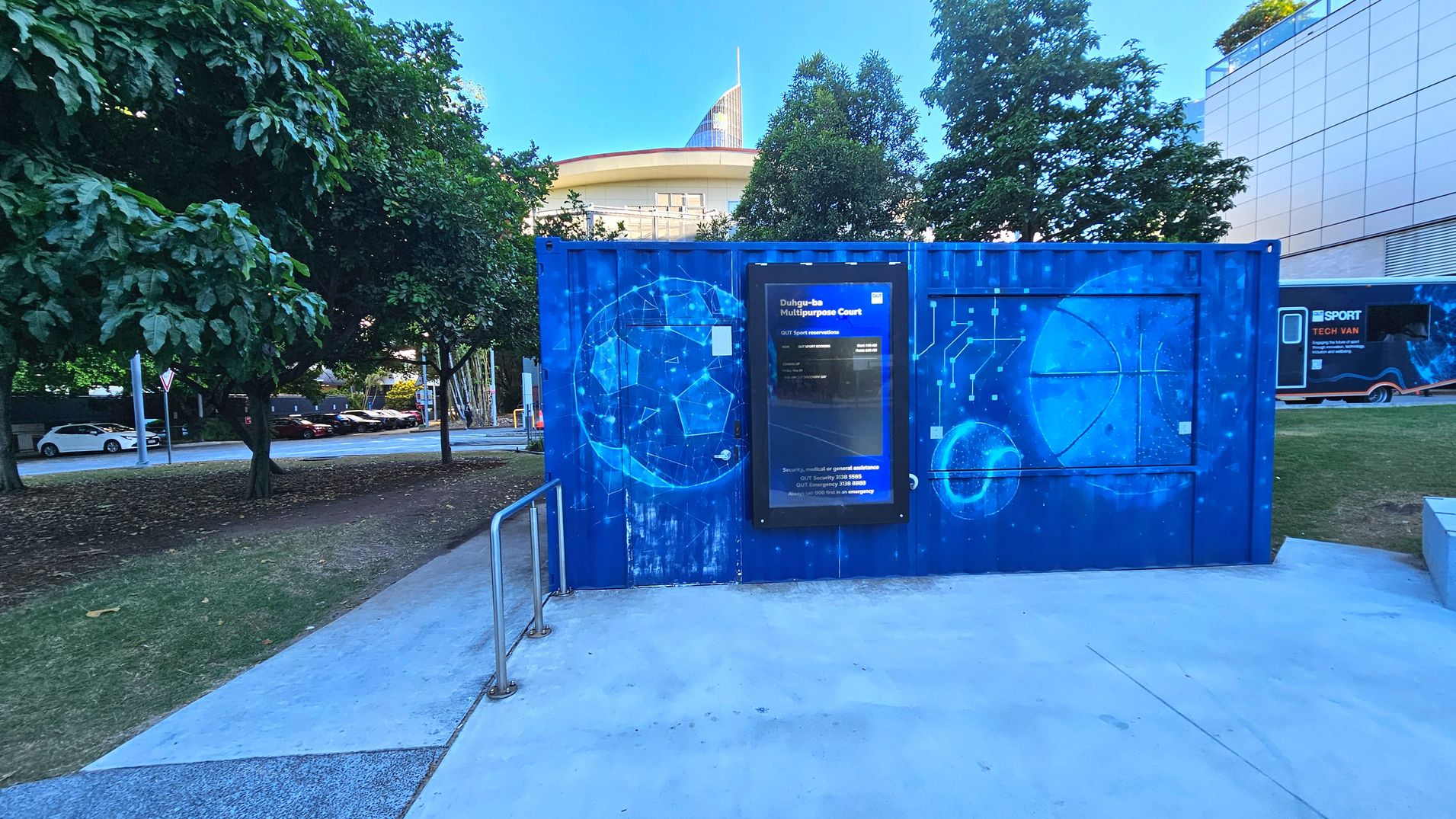 A vibrant blue container with geometric and basketball-themed artwork stands prominently. It features a digital sign reading 'Duhig-aba Multipurpose Court' with contact information. The container is situated on a concrete path with metal railings, surrounded by grass and trees. A modern building with a curved roof is visible in the background, and a tech van is parked nearby.