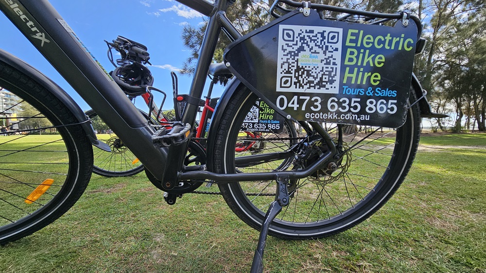 On the left, there is a close-up view of the rear wheel and frame of an electric bike. The middle features a QR code and a sign attached to the bike, displaying contact information for electric bike hire, including a phone number and website. On the right, another bike is partially visible. The background consists of a grassy park area with trees.