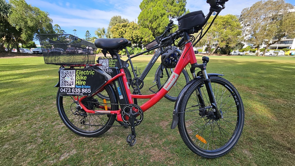 On the left, a red electric bike with a black basket on the rear and a sign displaying "Electric Bike Hire" along with a phone number and QR code. In the middle, the bike's frame and battery are visible. On the right, a grey electric bike is partially visible. The background consists of a grassy park area with trees and some parked cars.