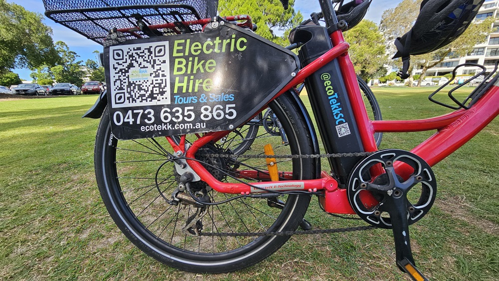 On the left, a red electric bike with a large black sign attached to the rear wheel, displaying "Electric Bike Hire," a QR code, a phone number, and a website. In the middle, the bike's frame, battery, and part of the pedal are visible. On the right, another red electric bike is partially visible. The background consists of a grassy park area with trees and parked cars.