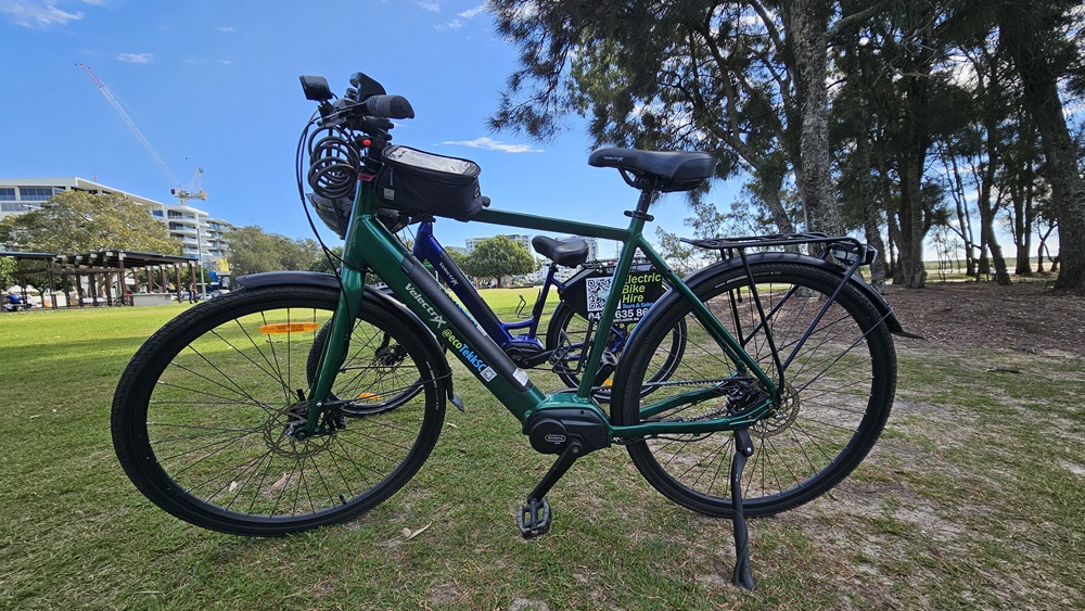 On the left, a dark blue electric bike with a small black bag attached to the top tube. On the right, a green electric bike with a similar design, featuring a black rear rack. In the middle, the green bike's frame and pedals are clearly visible. The background includes a grassy park area with trees, a pavilion, and a distant building.