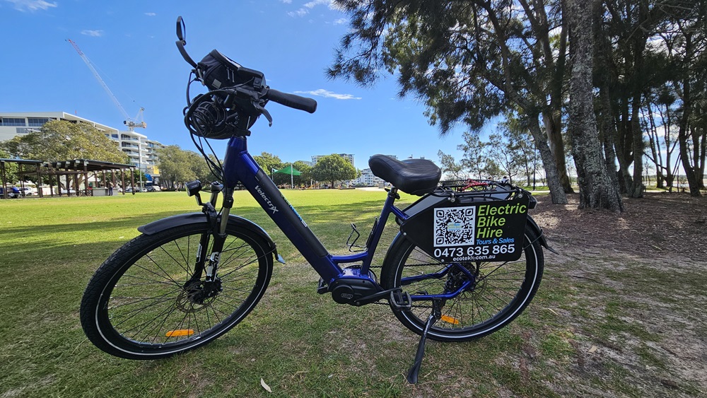 On the left, a dark blue electric bike with a black handlebar bag and a digital display. In the middle, the bike's frame, battery, and pedals are visible. On the right, the rear wheel has a sign attached, displaying "Electric Bike Hire," a QR code, and a phone number. The background includes a grassy park area with trees, a pavilion, and a distant building.