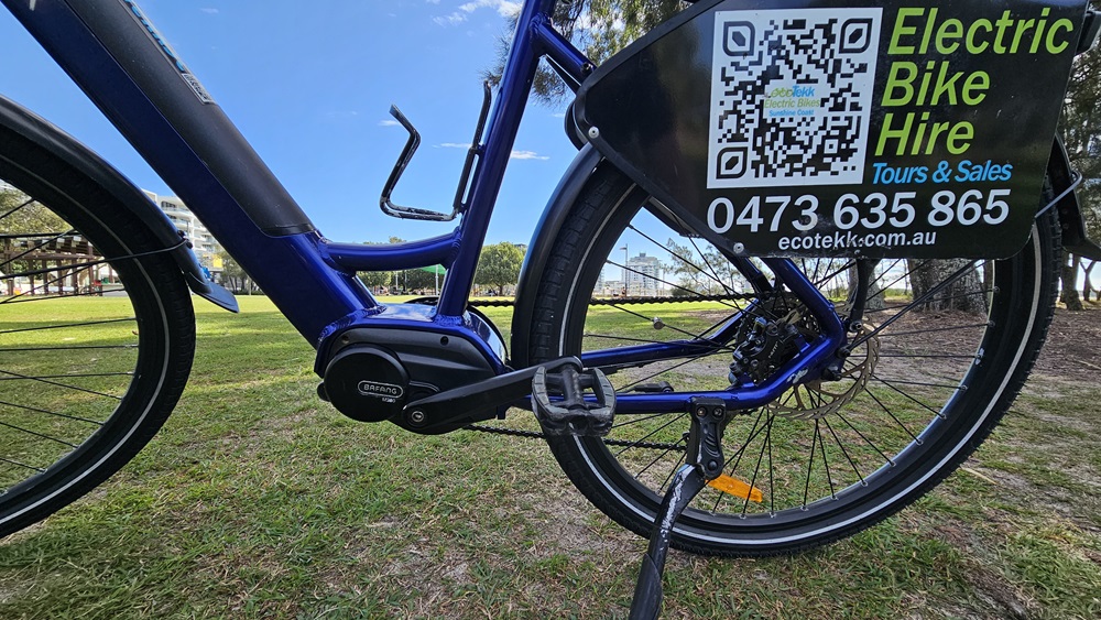 On the left, the frame and battery of a blue electric bike are visible. In the middle, the bike's pedal and chain are prominent. On the right, the rear wheel has a sign attached, displaying a QR code, "Electric Bike Hire," and a phone number. The background includes a grassy park area with trees and a distant building.