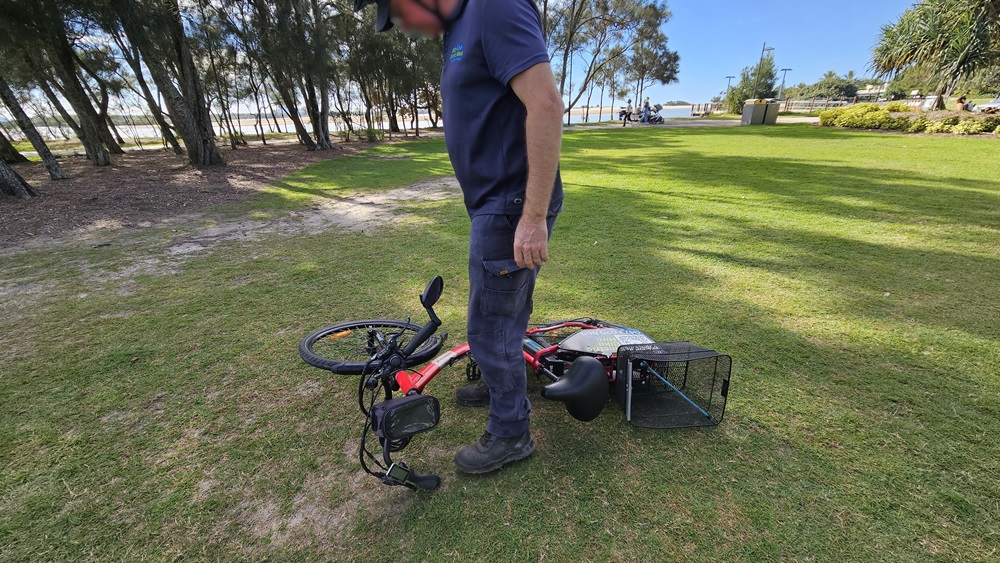 On the left, a man in a navy blue outfit is standing on grass. In the middle, a red electric bike lies on its side with the handlebars and front wheel facing left. On the right, the bike's rear wheel and a black basket are visible. The background includes a grassy park area with trees, a pathway, and people in the distance.