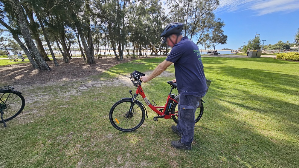 On the left, a black bike wheel is partially visible. In the middle, a person wearing a helmet and dark clothing is standing next to a red electric bike, holding the handlebars. On the right, there is a grassy park area with trees and a pathway leading to a distant building.