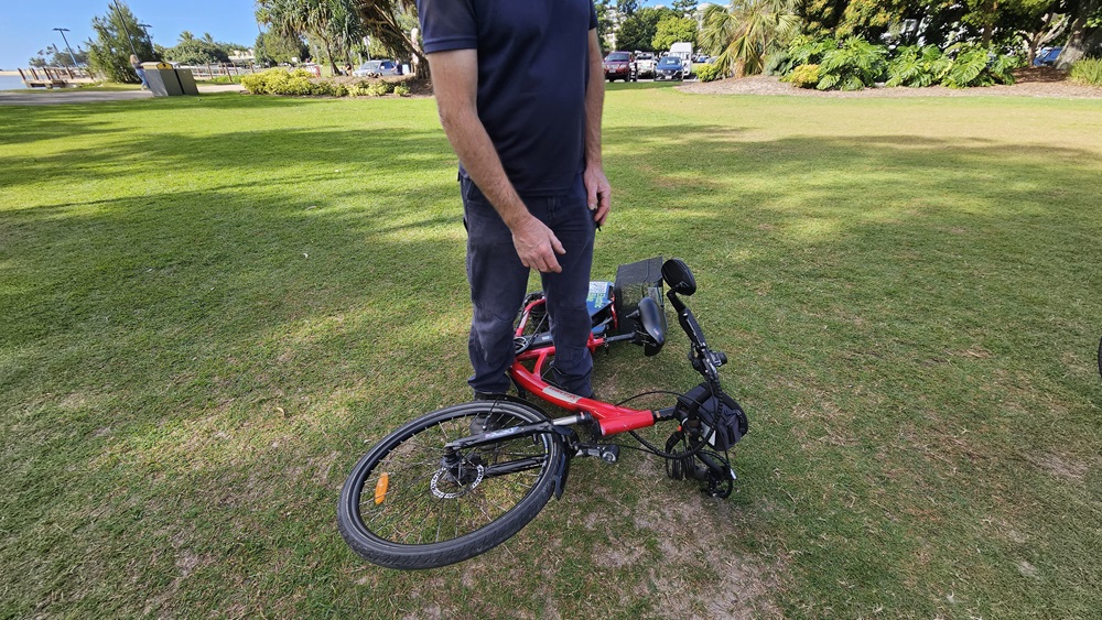 On the left, a man in a navy blue shirt and grey trousers is standing on grass. In the middle, a red electric bike is lying on its side with the handlebars and front wheel facing left. On the right, the bike's rear wheel and a black basket are visible. The background includes a grassy park area with trees, bushes, and parked cars.