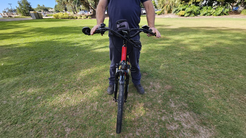 On the left, a patch of green grass with some bushes in the distance. In the middle, a person wearing dark clothing is holding the handlebars of a red and black electric bike, standing on grass. The bike's front wheel is facing forward. On the right, more green grass and trees are visible. The background includes a grassy park area with trees and some distant structures.