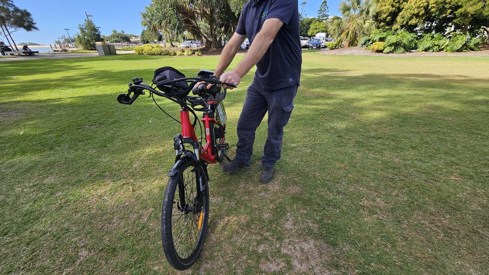On the left, a patch of green grass with some bushes and trees in the distance. In the middle, a person wearing a dark shirt and trousers is holding the handlebars of a red and black electric bike, standing on grass. The bike's front wheel is facing forward. On the right, more green grass, trees, and parked cars are visible. The background includes a grassy park area with trees, bushes, and distant structures.