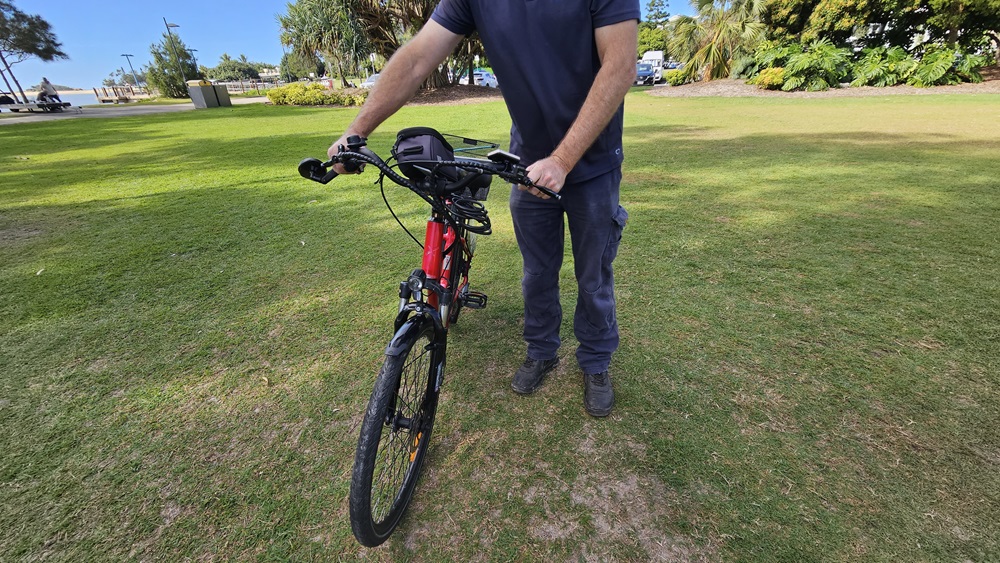 On the left, a grassy area with a few scattered trees and a distant building. In the middle, a person wearing a dark blue shirt and trousers is holding the handlebars of a red and black electric bike, standing on grass. The bike's front wheel is facing forward. On the right, more grass, trees, and a few parked cars are visible. The background includes a grassy park area with trees and bushes.
