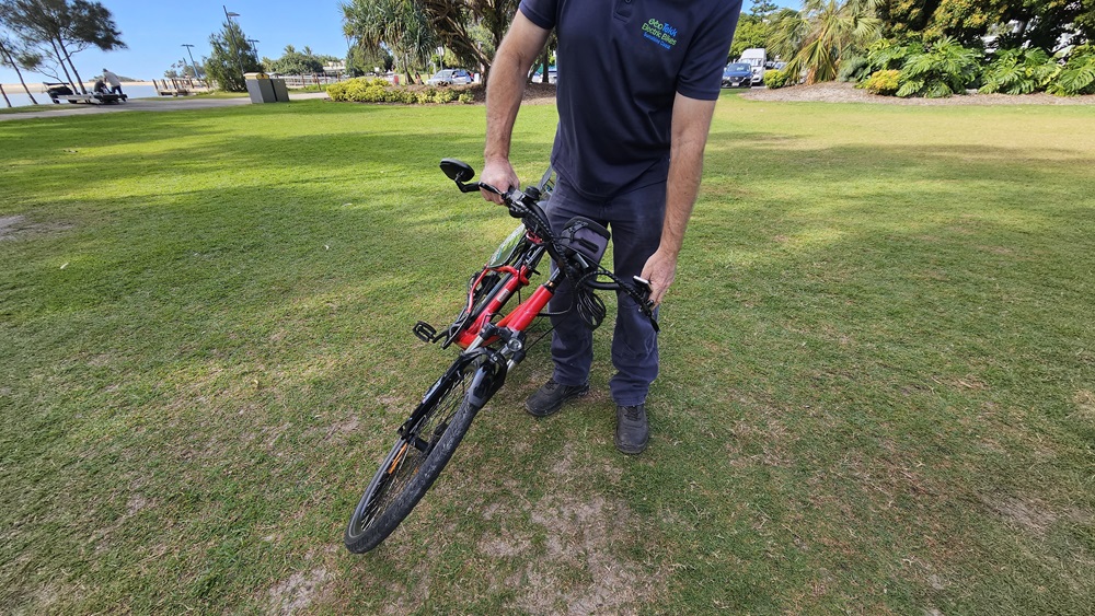 On the left, a grassy area with a few scattered trees and a distant pathway. In the middle, a person wearing a dark blue shirt and trousers is holding a red and black electric bike by the handlebars, lifting its front wheel off the ground. On the right, more grass, trees, and a few parked cars are visible. The background includes a grassy park area with trees and bushes.