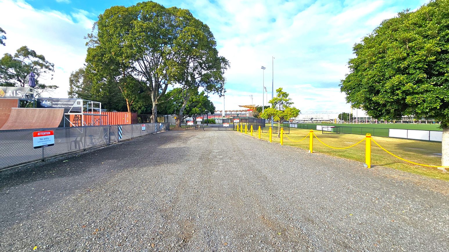 A gravel path runs through the centre, flanked by trees on both sides. On the left, there's a fenced area with a skate ramp and a sign that reads "Attention No entry." On the right, a grassy field is bordered by a yellow rope barrier. In the background, there are more trees and a sports field with floodlights. The sky is partly cloudy. A gravel path runs through the centre, flanked by trees on both sides. On the left, there's a fenced area with a skate ramp and a sign that reads "Attention No entry." On the right, a grassy field is bordered by a yellow rope barrier. In the background, there are more trees and a sports field with floodlights. The sky is partly cloudy.