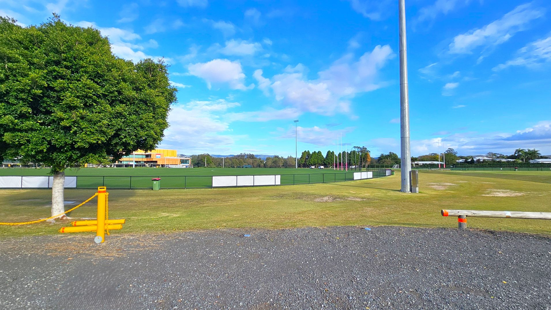 A grassy sports field with a tall floodlight pole on the right. In the foreground, there's a gravel path and a wooden barrier with orange markings. On the left, a large leafy tree stands next to a yellow chain barrier. In the background, a building with orange and yellow accents is visible, along with a line of trees and distant hills. A green rubbish bin is near the fence surrounding the field. A grassy sports field with a tall floodlight pole on the right. In the foreground, there's a gravel path and a wooden barrier with orange markings. On the left, a large leafy tree stands next to a yellow chain barrier. In the background, a building with orange and yellow accents is visible, along with a line of trees and distant hills. A green rubbish bin is near the fence surrounding the field.