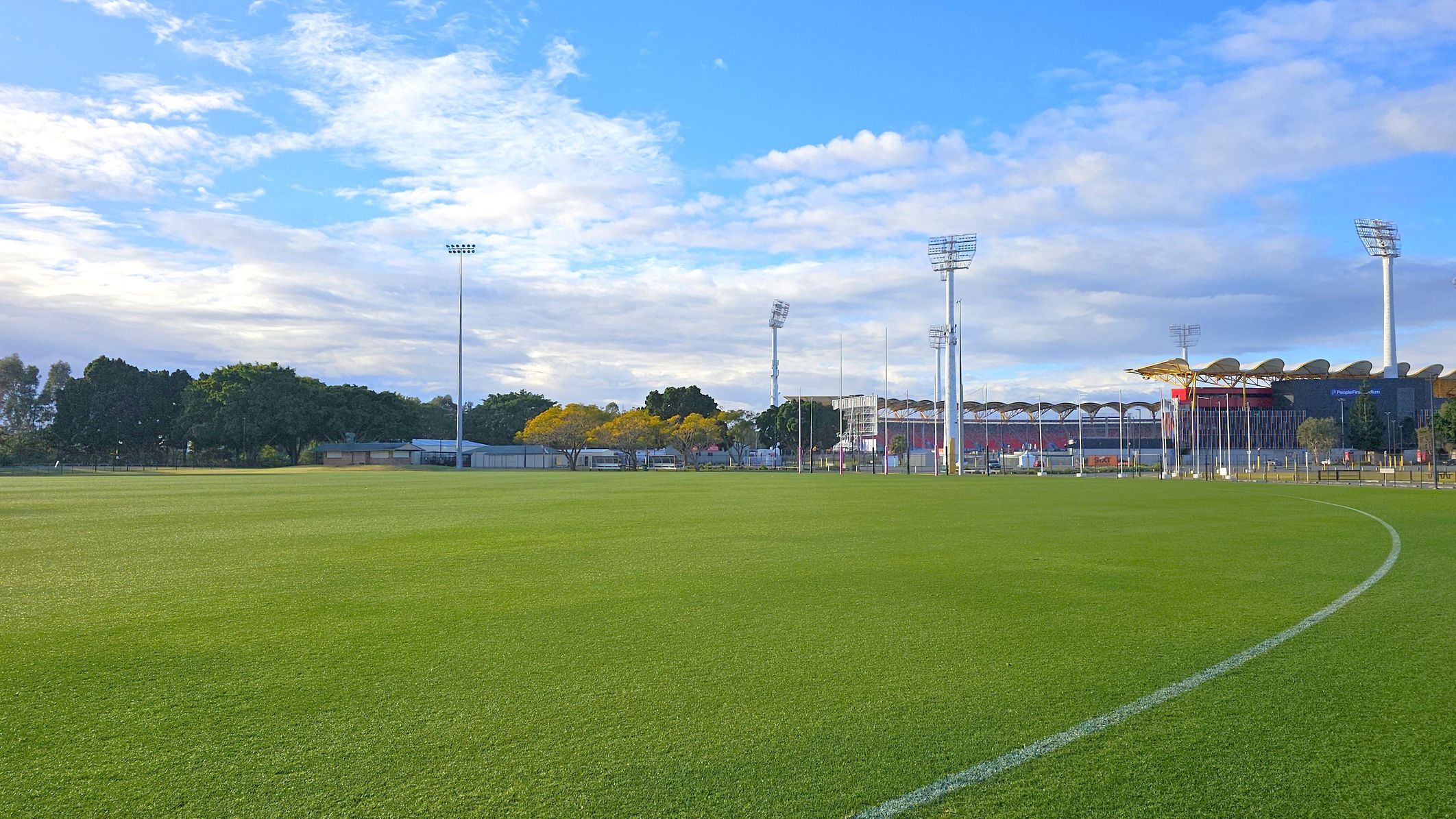 A large, well-maintained grassy sports field with a white boundary line curving across the foreground. In the background, there are tall floodlights and a stadium with a distinctive roof design. Trees with green and yellow foliage are visible to the left, along with some small buildings. The sky is partly cloudy with patches of blue. A large, well-maintained grassy sports field with a white boundary line curving across the foreground. In the background, there are tall floodlights and a stadium with a distinctive roof design. Trees with green and yellow foliage are visible to the left, along with some small buildings. The sky is partly cloudy with patches of blue.
