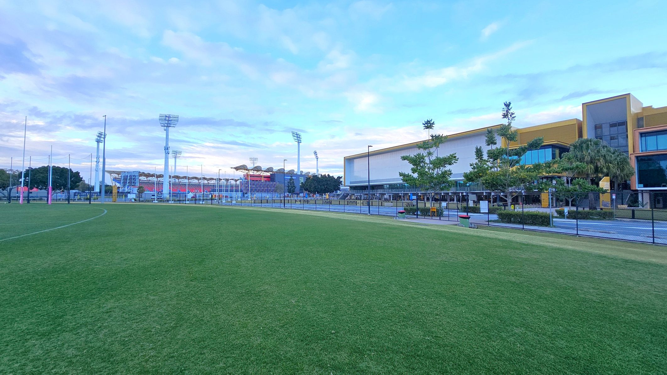 A grassy field in the foreground with a sports stadium in the background. The stadium has tall floodlights and a seating area with a red and white colour scheme. On the right, there is a modern building with large windows and a yellow and grey facade, surrounded by trees and a fence. The sky is partly cloudy. A grassy field in the foreground with a sports stadium in the background. The stadium has tall floodlights and a seating area with a red and white colour scheme. On the right, there is a modern building with large windows and a yellow and grey facade, surrounded by trees and a fence. The sky is partly cloudy.