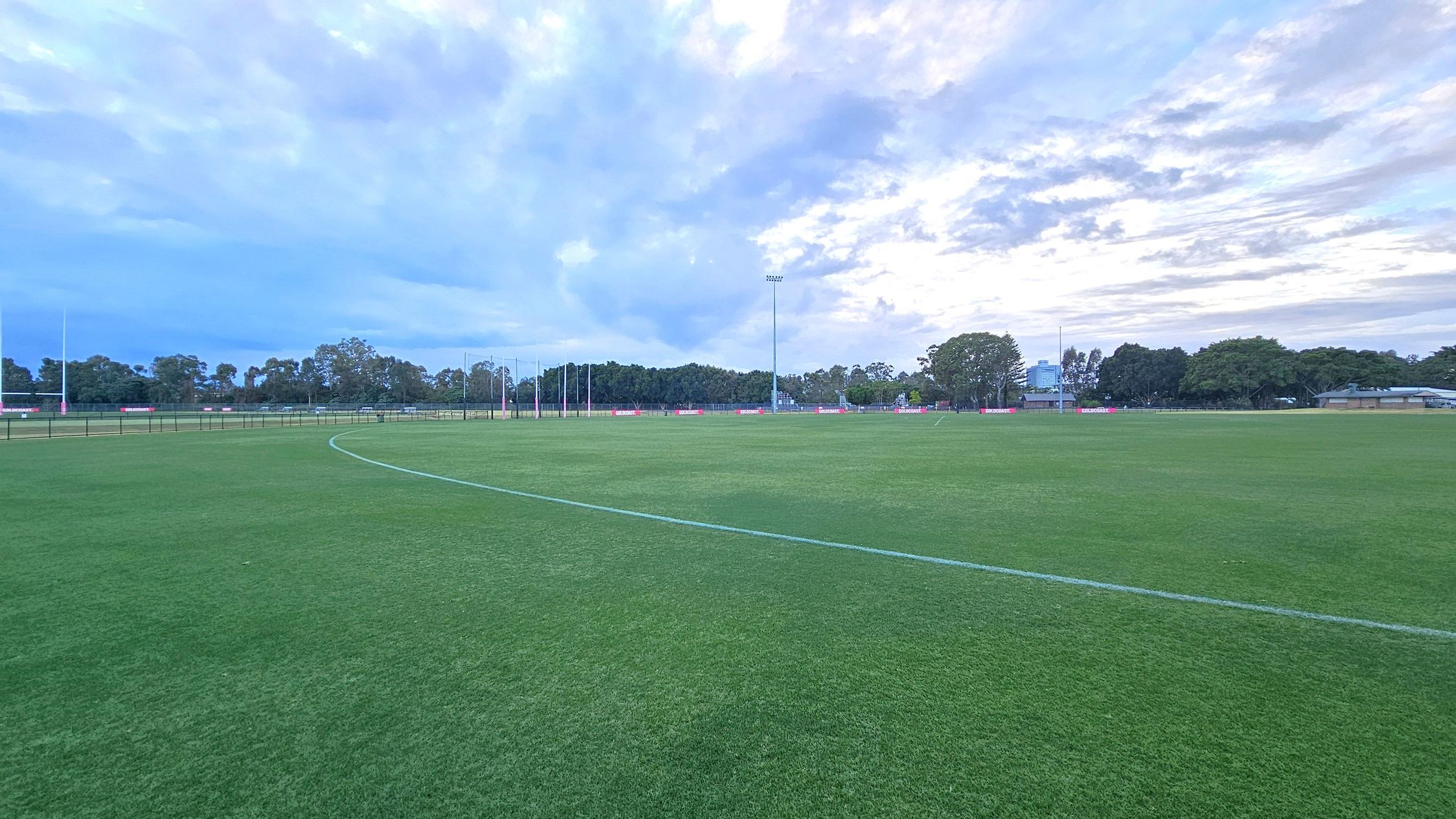 A wide, open sports field with well-maintained green grass. In the background, there are several tall light poles and a row of trees lining the perimeter. To the right, a few buildings are partially visible among the trees. The sky is overcast with a mix of blue and grey clouds. A wide, open sports field with well-maintained green grass. In the background, there are several tall light poles and a row of trees lining the perimeter. To the right, a few buildings are partially visible among the trees. The sky is overcast with a mix of blue and grey clouds.