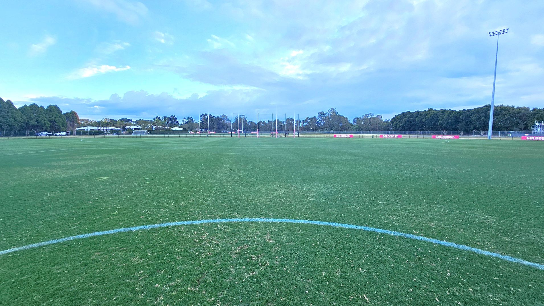 A large, open sports field with well-maintained grass. In the centre, there are goalposts in the distance. The foreground shows a curved white line on the grass. To the right, a tall floodlight pole stands near the edge of the field. The background features a line of trees and some buildings, with a few parked cars visible. Red signs with "GOLDCOAST" are placed along the perimeter fence. A large, open sports field with well-maintained grass. In the centre, there are goalposts in the distance. The foreground shows a curved white line on the grass. To the right, a tall floodlight pole stands near the edge of the field. The background features a line of trees and some buildings, with a few parked cars visible. Red signs with "GOLDCOAST" are placed along the perimeter fence.