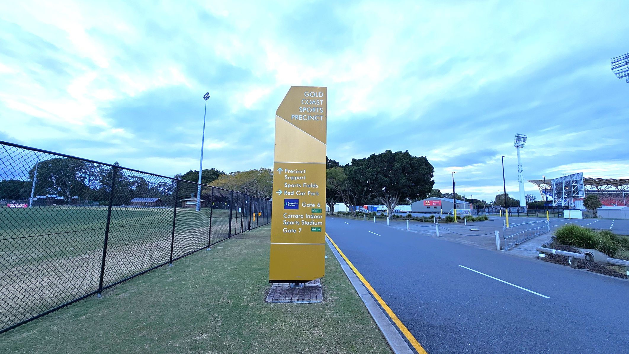 A tall, yellow sign stands on a grassy area, displaying directions for the Gold Coast Sports Precinct. To the left, a chain-link fence encloses a sports field with trees in the background. On the right, a paved road runs alongside the sign, leading towards a stadium with visible floodlights and seating. The area is bordered by trees and some low buildings. A tall, yellow sign stands on a grassy area, displaying directions for the Gold Coast Sports Precinct. To the left, a chain-link fence encloses a sports field with trees in the background. On the right, a paved road runs alongside the sign, leading towards a stadium with visible floodlights and seating. The area is bordered by trees and some low buildings.